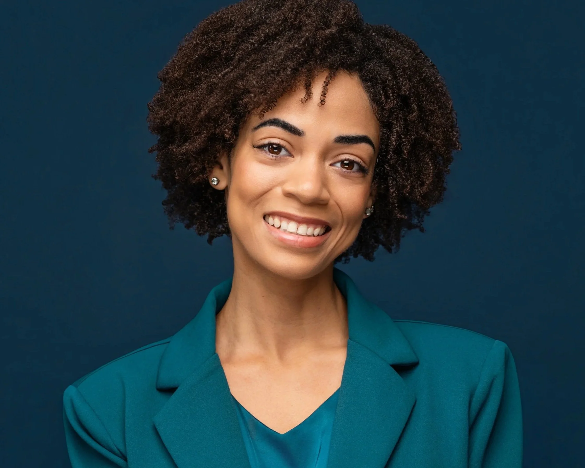 A smiling light skinned black woman with short curly hair wearing a teal blazer and earrings against a dark  blue background.