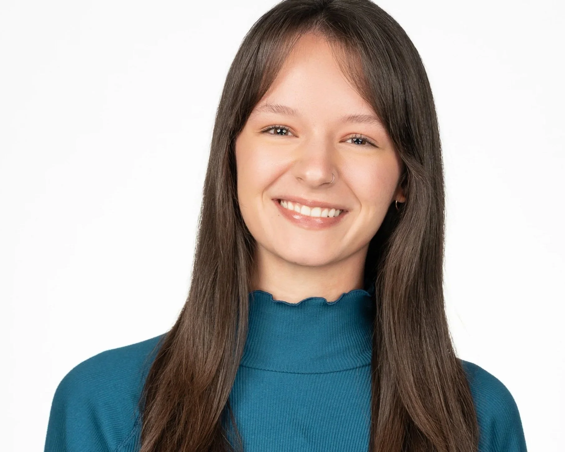 Smiling young woman with long brown hair, wearing a blue top and a small nose ring, against a white background.