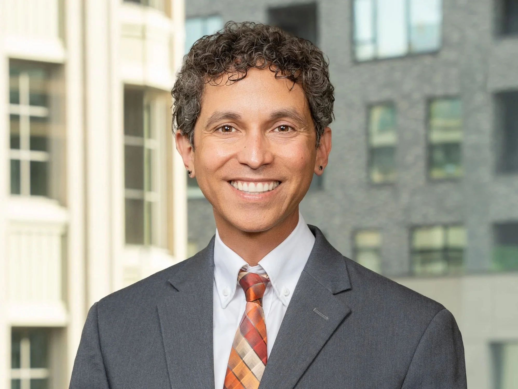 A smiling man in a business suit standing outdoors with buildings in the background.