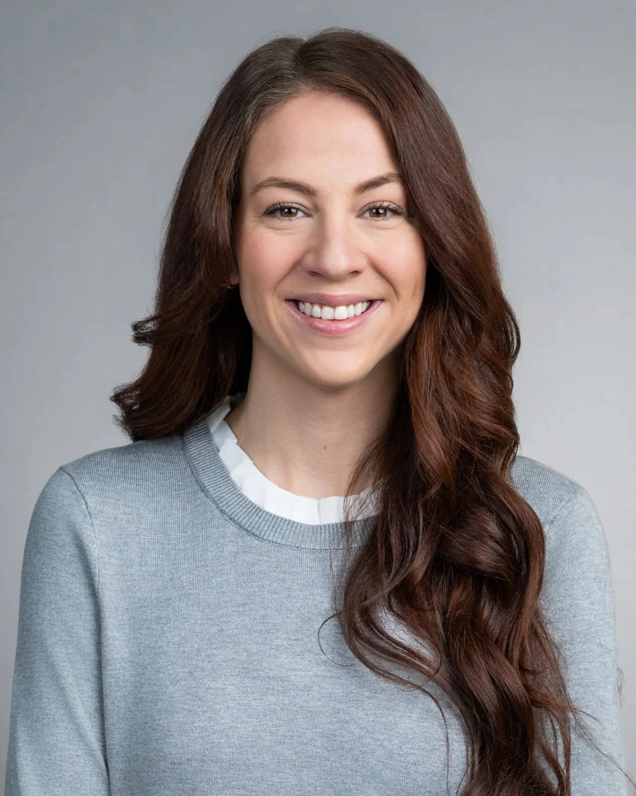 Headshot of a woman with long, wavy auburn hair, smiling, wearing a light gray sweater and a white shirt with a ruffled collar, against a plain gray background.