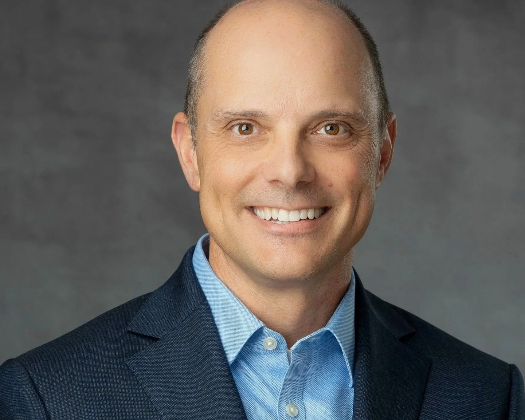 A man in a navy suit and light blue dress shirt smiling, with short hair and a light complexion, standing against a dark gray background.
