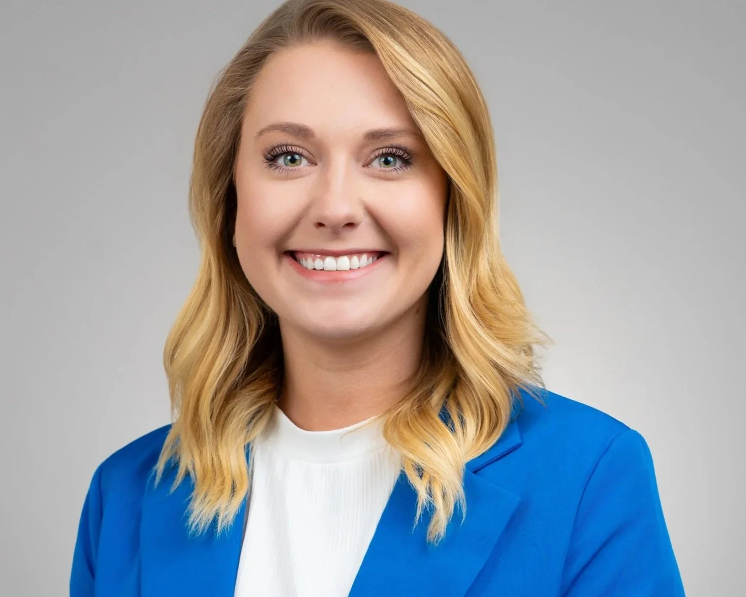 A woman with blonde, wavy hair smiling at the camera, wearing a blue blazer and white top against a plain gray background.