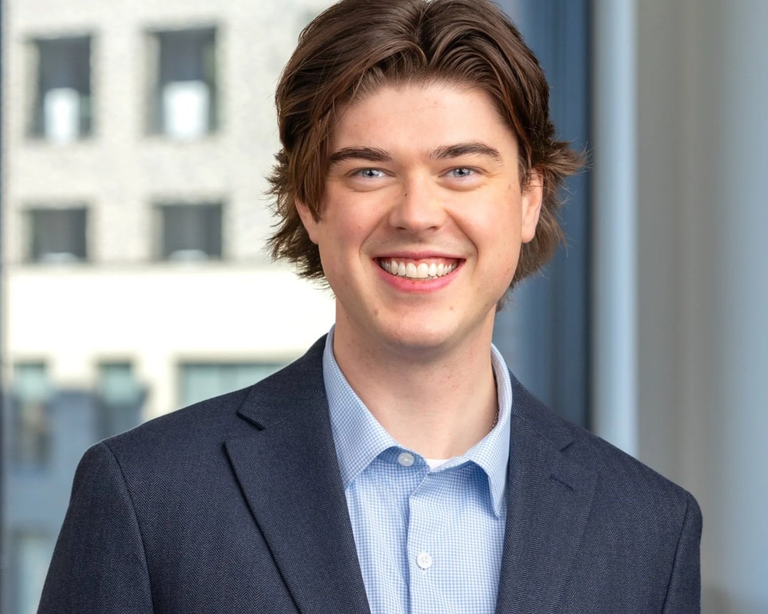A young man with brown hair, blue eyes, and a wide smile, dressed in a dark blazer and light blue collared shirt, standing in front of a large window overlooking a cityscape.