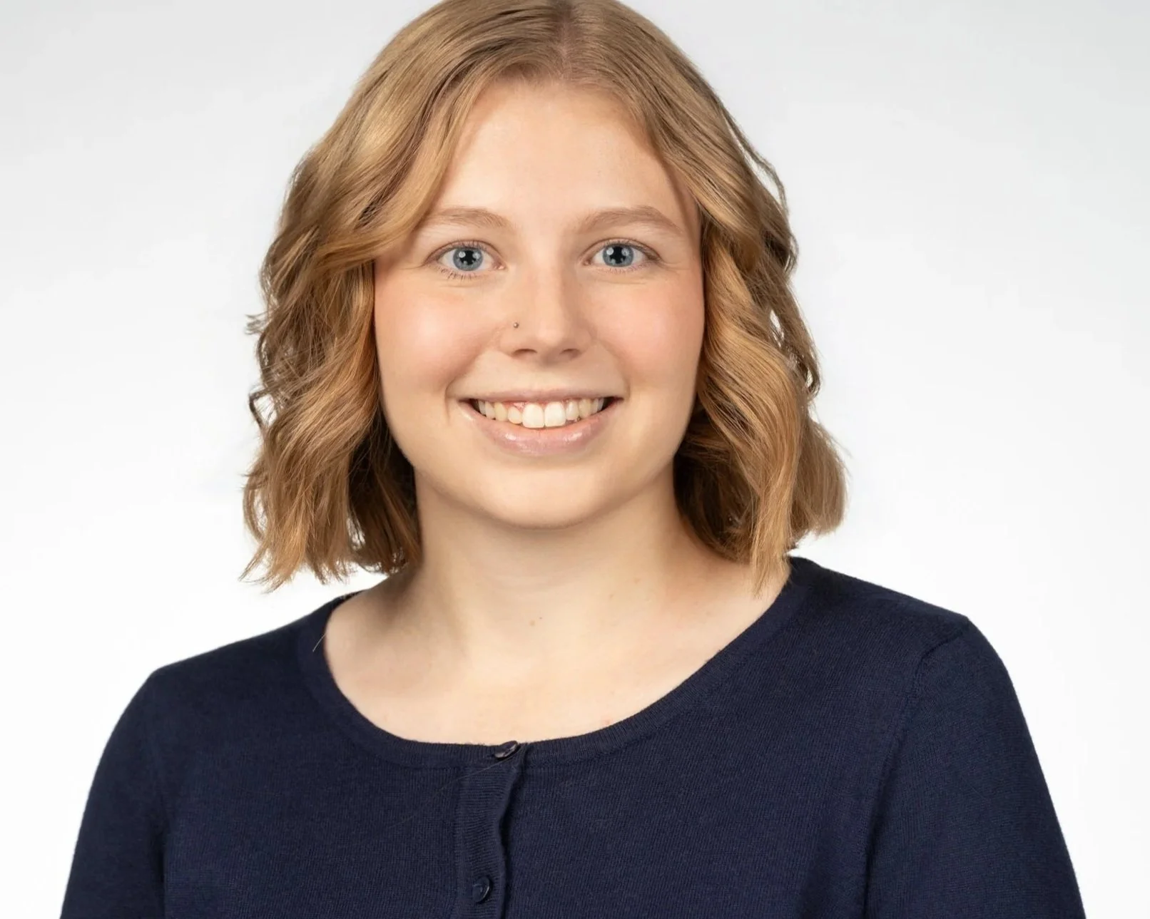 Portrait of a young woman with shoulder-length wavy red hair, blue eyes, wearing a navy blue top with a round neckline, smiling against a plain white background.