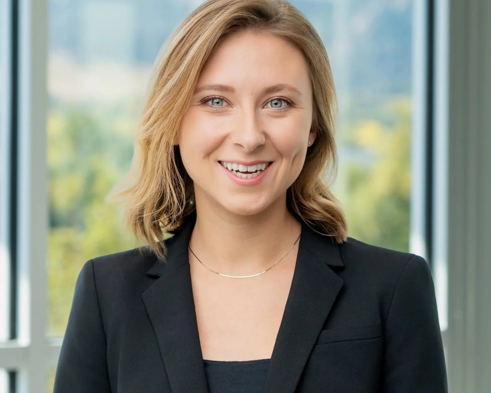 A smiling young woman with shoulder-length blonde hair, wearing a black blazer and a necklace, standing in front of large windows with a view of greenery outside.