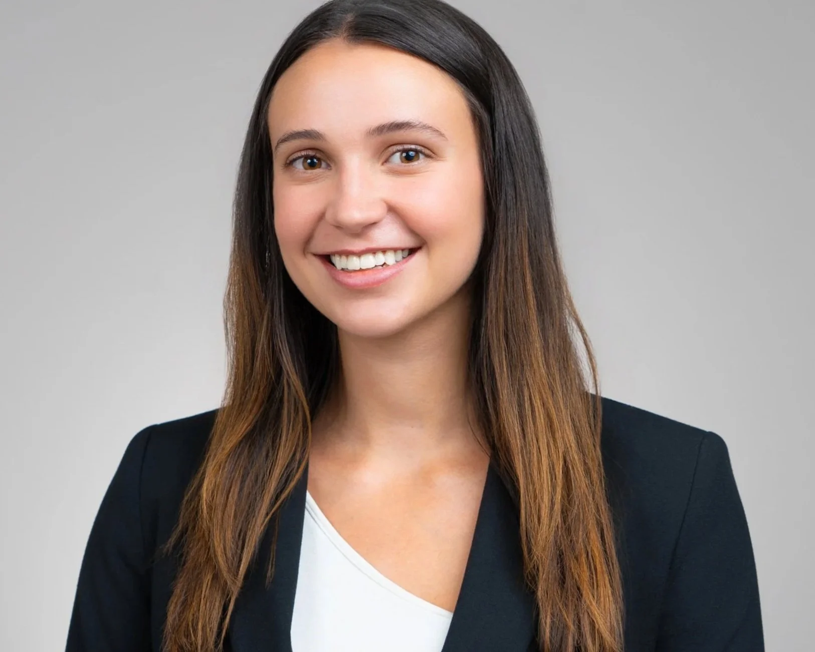 A young woman with long brown hair smiling, wearing a black blazer over a white top, against a plain gray background.