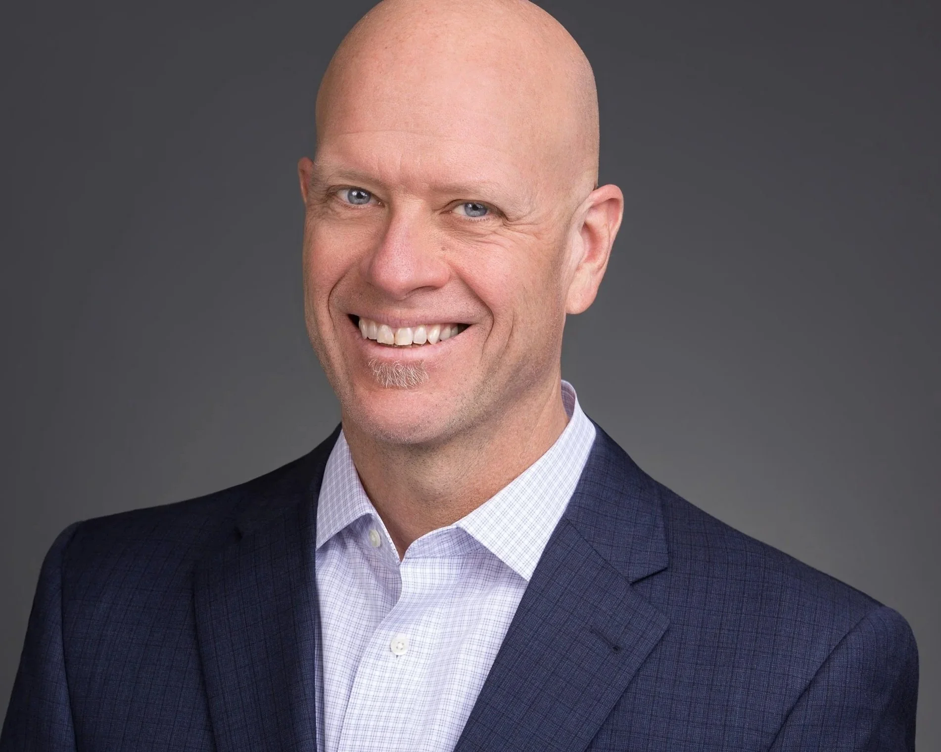 A smiling, bald man with blue eyes, wearing a dark blue suit jacket over a light button-up shirt, against a gray background.