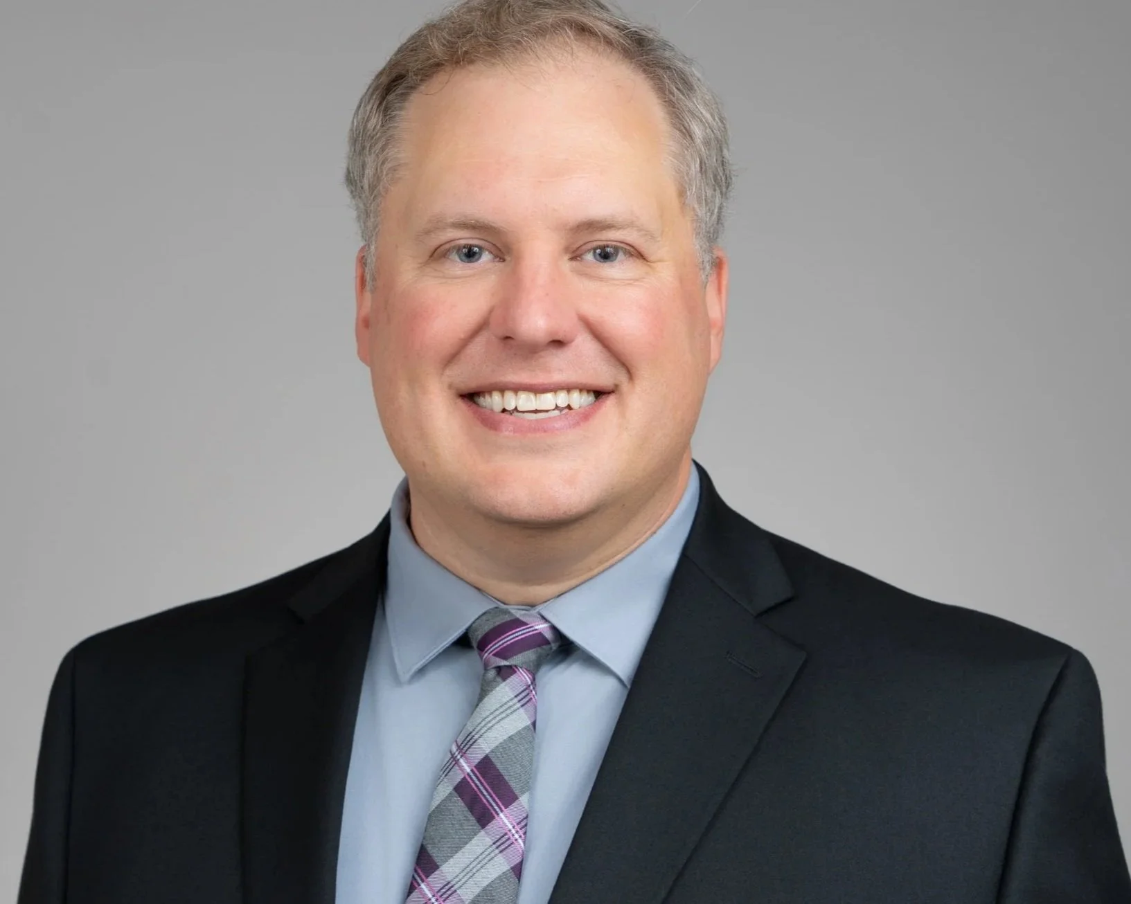 A smiling man with blond hair wearing a black suit, blue shirt, and a plaid purple and gray tie against a plain gray background.