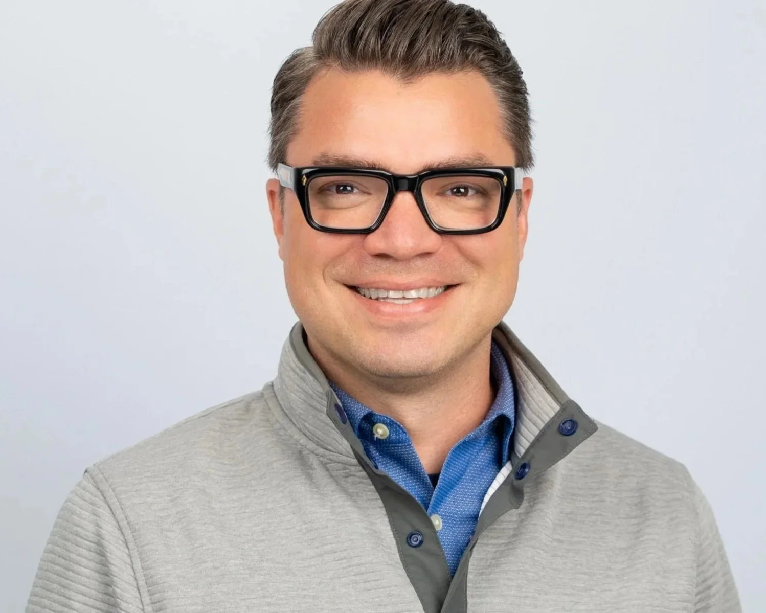Portrait of a smiling man with glasses, wearing a light gray zip-up jacket over a blue collared shirt, against a plain gray background.
