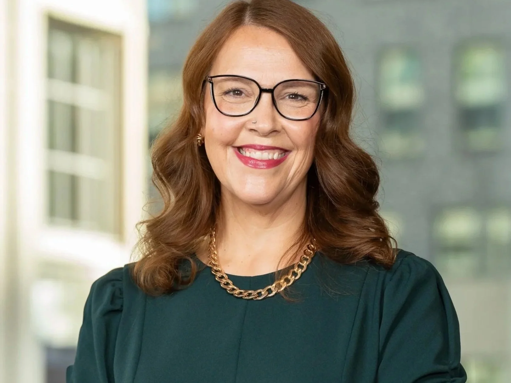 Portrait of a smiling woman with shoulder-length red hair, glasses, wearing a dark green blouse, a gold chain necklace, and small earrings, standing indoors with large windows in the background.