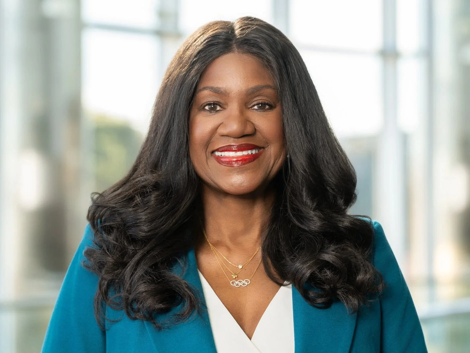A smiling woman with long black hair, wearing a teal blazer and gold jewelry, standing in a modern, well-lit office building.