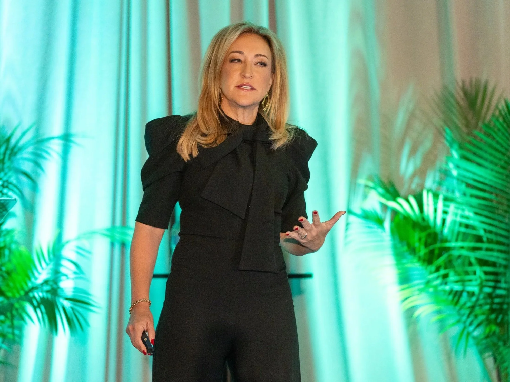 A woman with blonde hair wearing a black outfit, standing on a stage with green lighting and plants in the background, gesturing with her right hand while speaking.