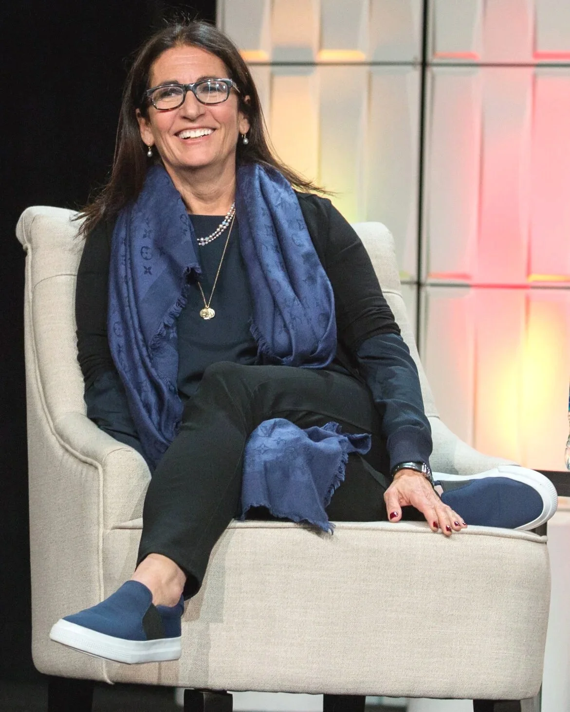 Makeup mogul Bobbi Brown sitting on a beige armchair, smiling, wearing glasses, a black top, a blue scarf, black pants, and blue slip-on shoes, with a colorful background
