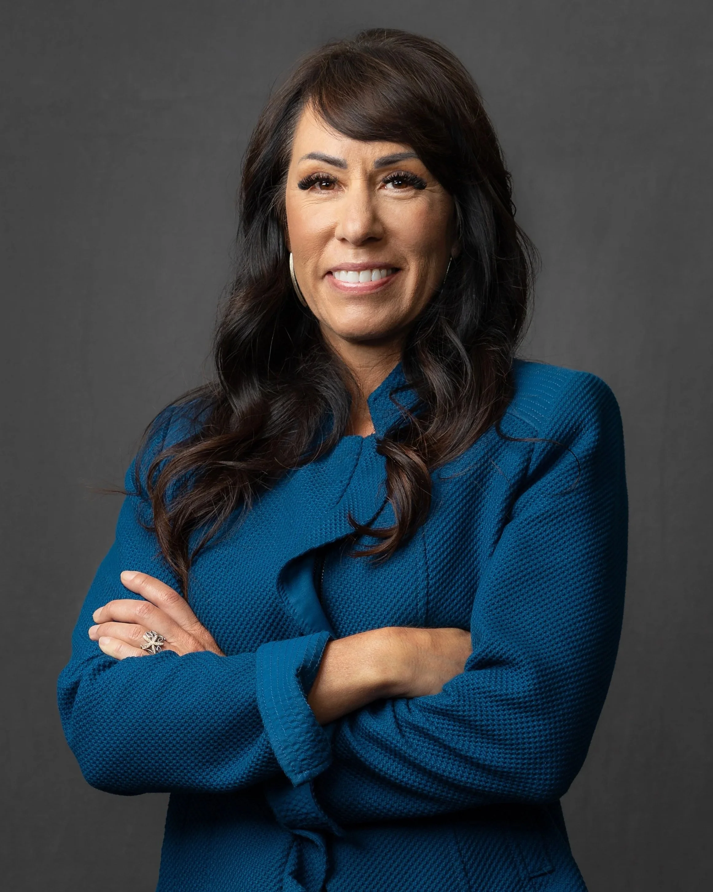 A woman with dark brown hair wearing a blue blazer and black pants, standing with her arms crossed against a dark gray background.