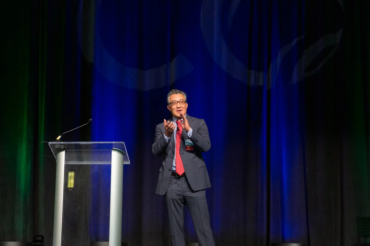 A man in a gray suit, red tie, and glasses speaking at a podium on a stage with blue and green curtains.