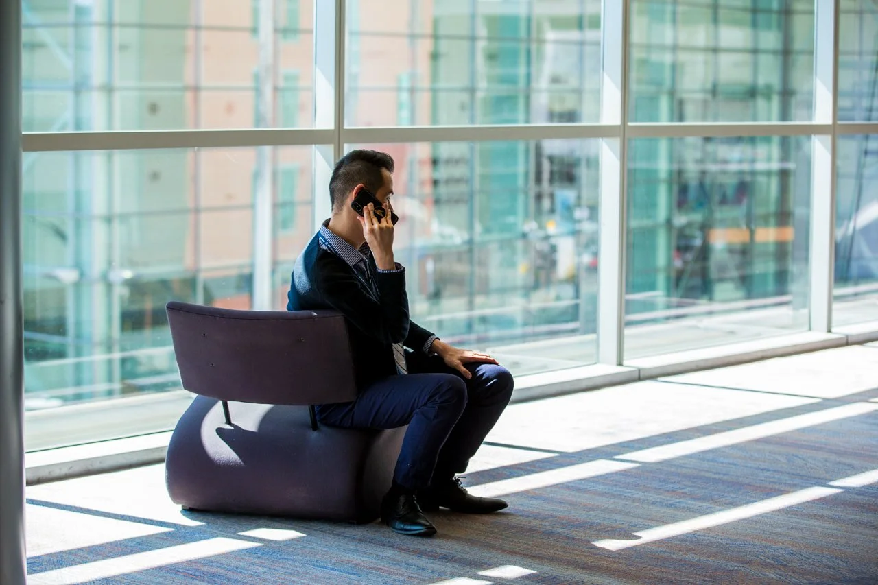 A man sitting on a modern lounge chair in a bright, glass-walled convention center, talking on a cellphone.