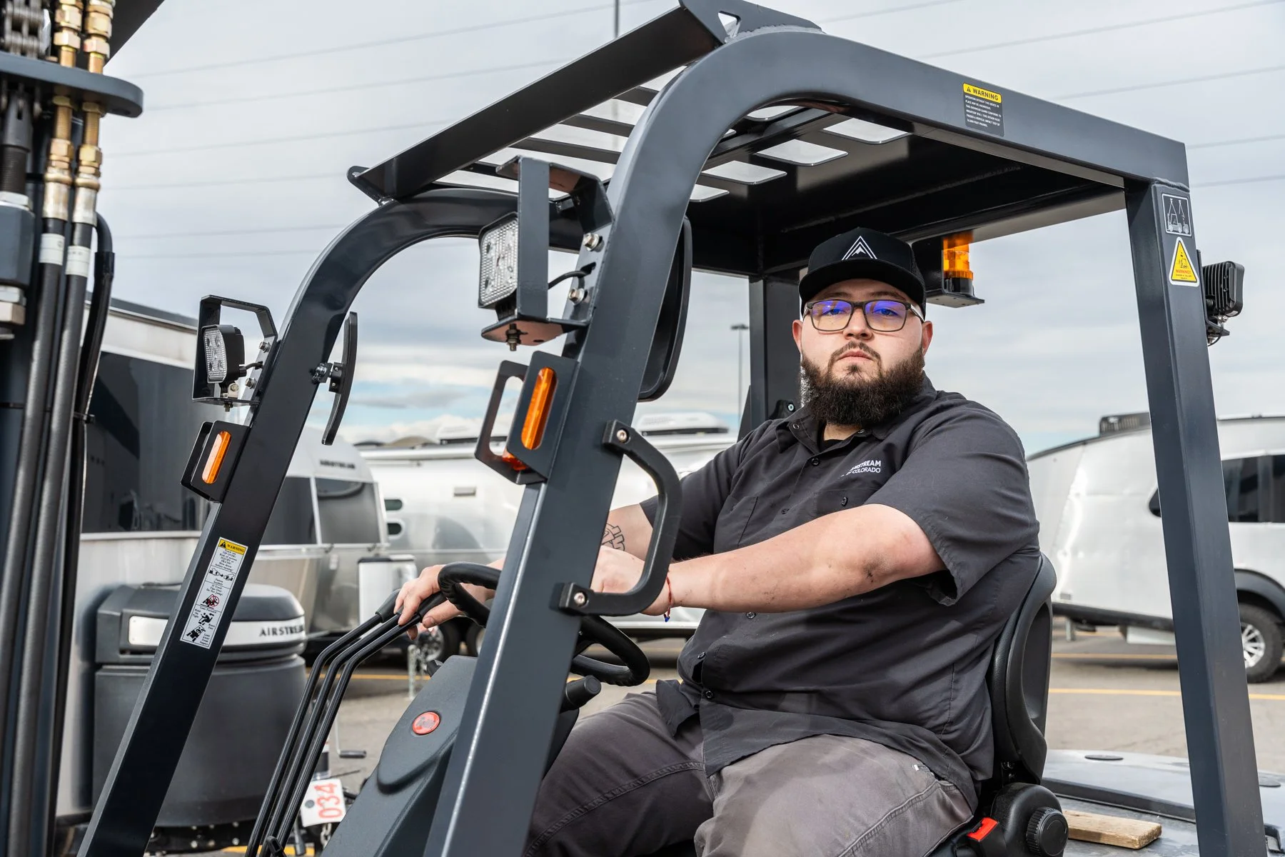 A man with a beard and glasses sitting on a forklift, wearing a black cap and black work shirt, in an outdoor parking lot with trailers and RVs in the background.