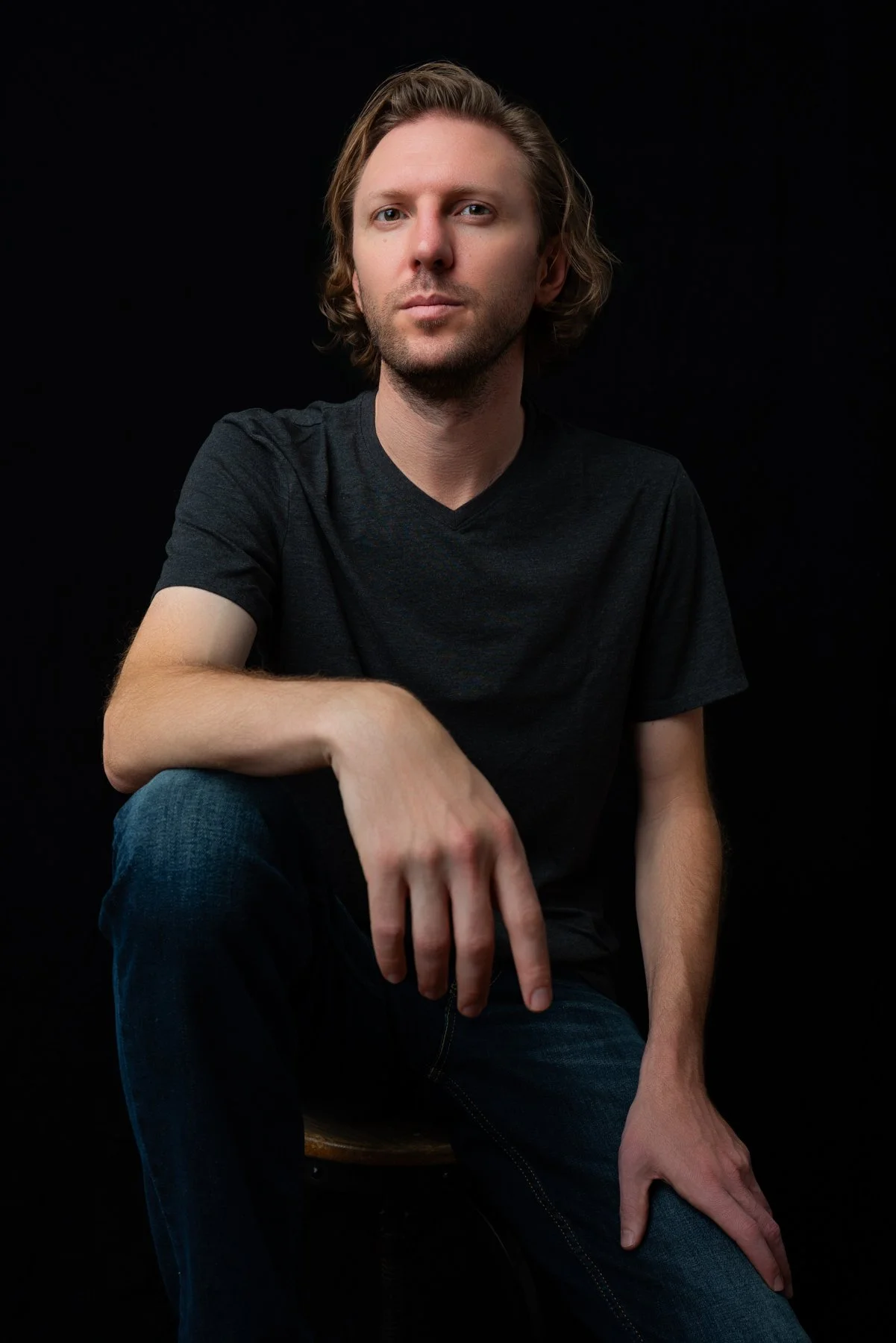 A portrait of a young man with shoulder-length blond hair, wearing a black t-shirt, sitting against a black background.