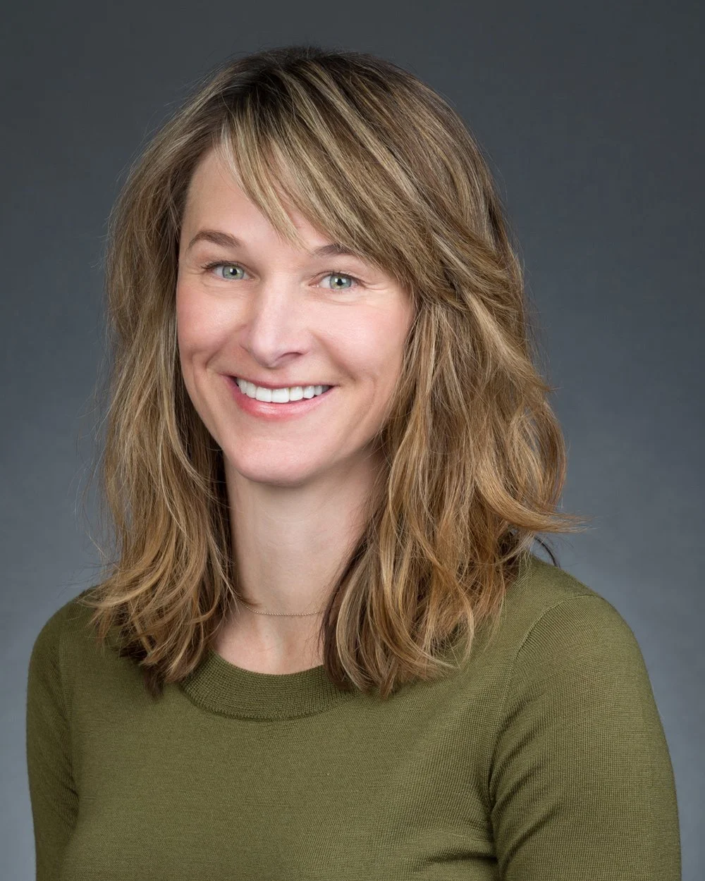 A woman with light skin, shoulder-length wavy light brown hair, and blue eyes smiling in front of a dark gray background. She is wearing a green top.