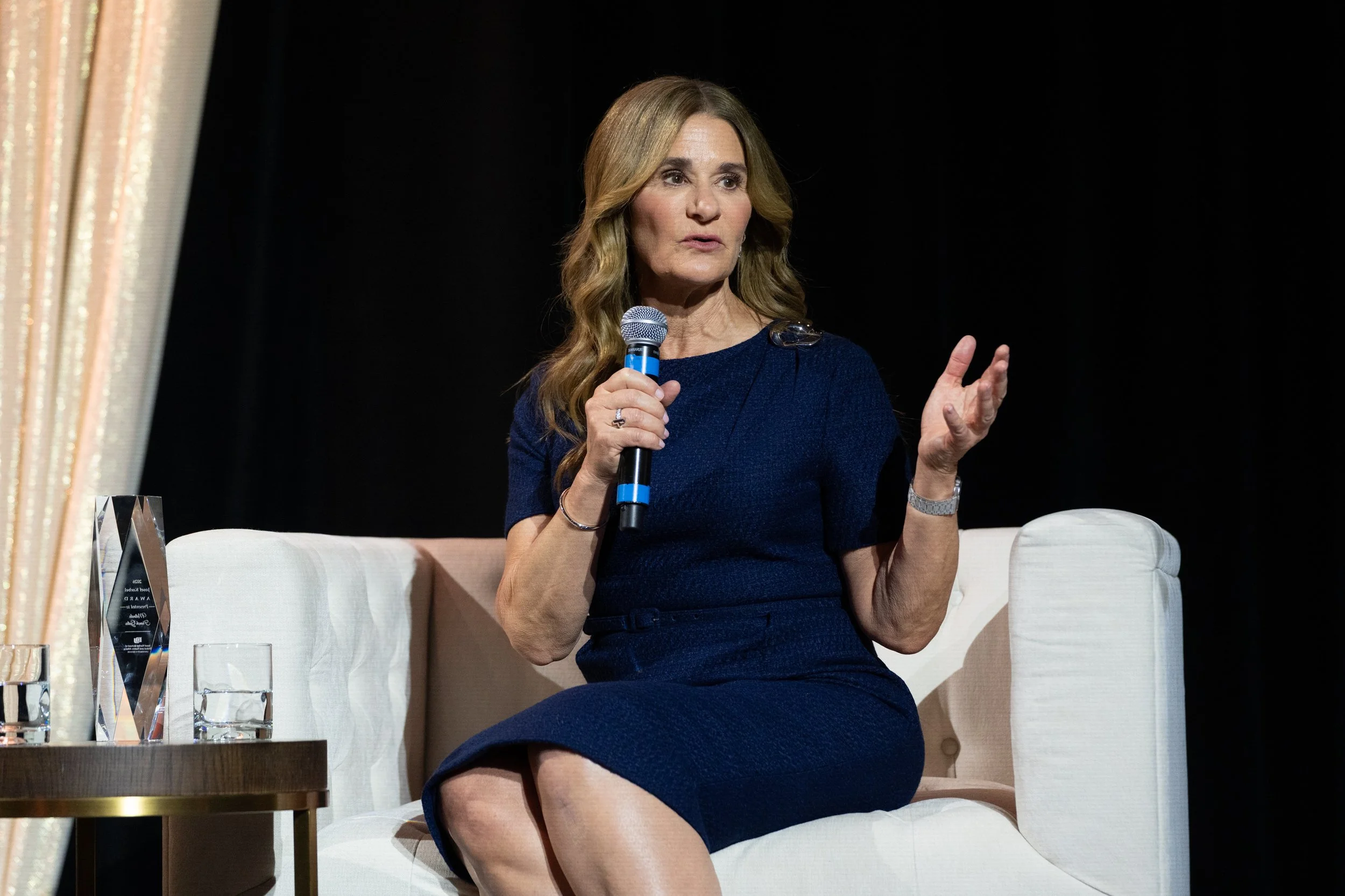 Melinda French Gates seated on stage, wearing a dark blue dress, sitting on a white sofa, holding a microphone, and gesturing with her left hand during a discussion or presentation.