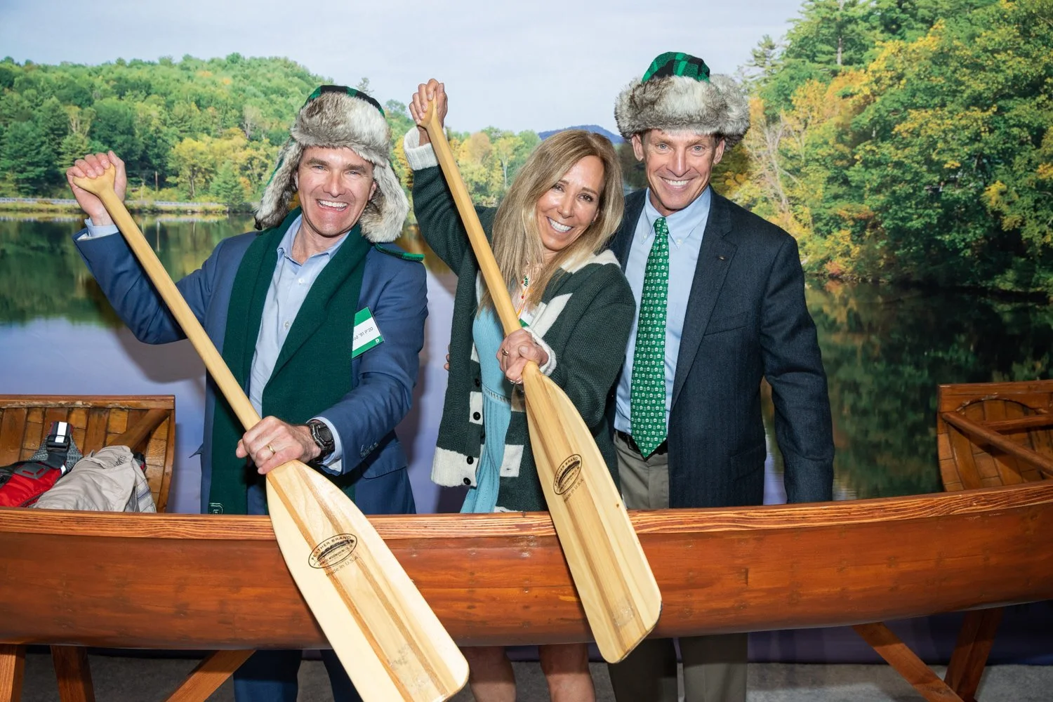 Three people in outdoor gear holding paddles and smiling, standing behind a wooden boat with a nature scene in the background.
