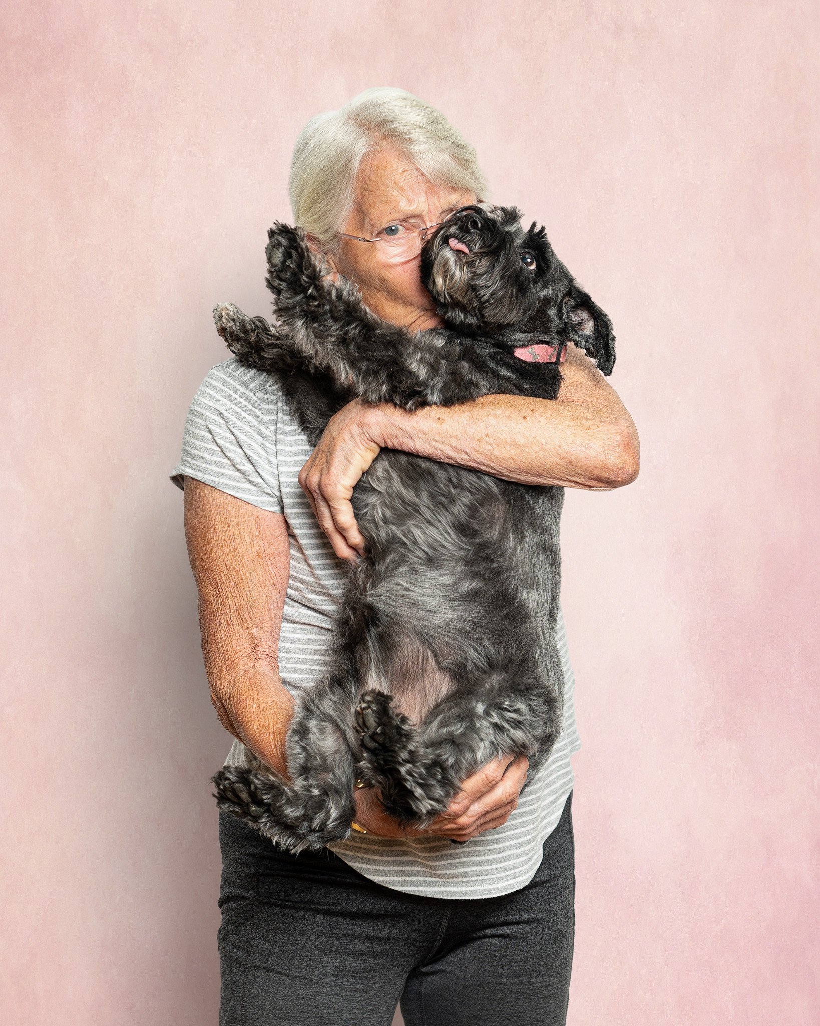 An elderly woman holding a large black and gray puppy in her arms, against a pink background.