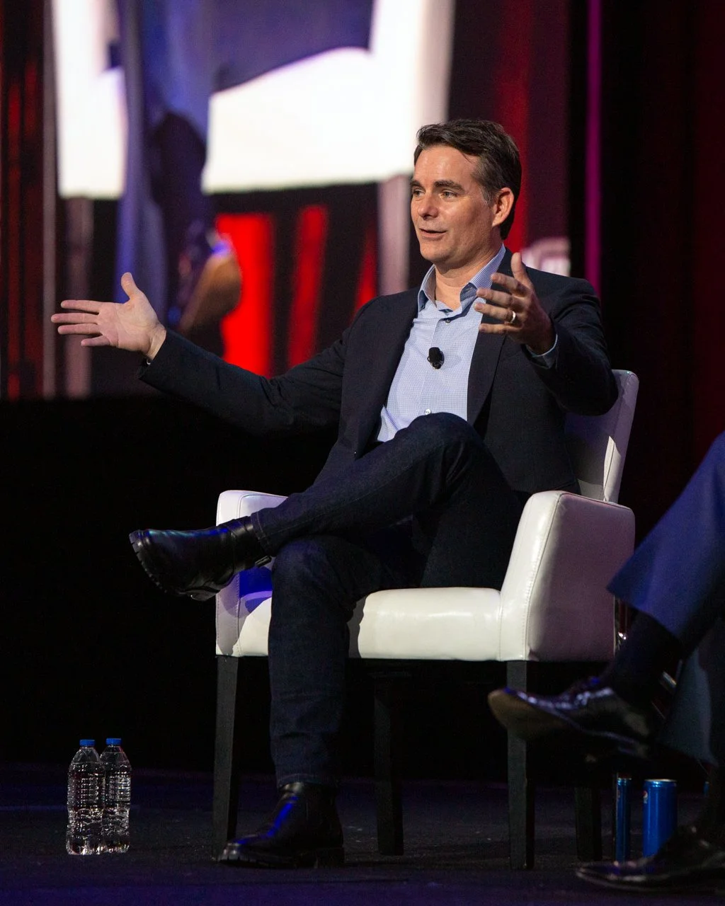 NASCAR Champion Jeff Gordon seated on a white chair during a presentation or panel discussion, gesturing with his hands, with a large screen behind him displaying part of a person in a suit.