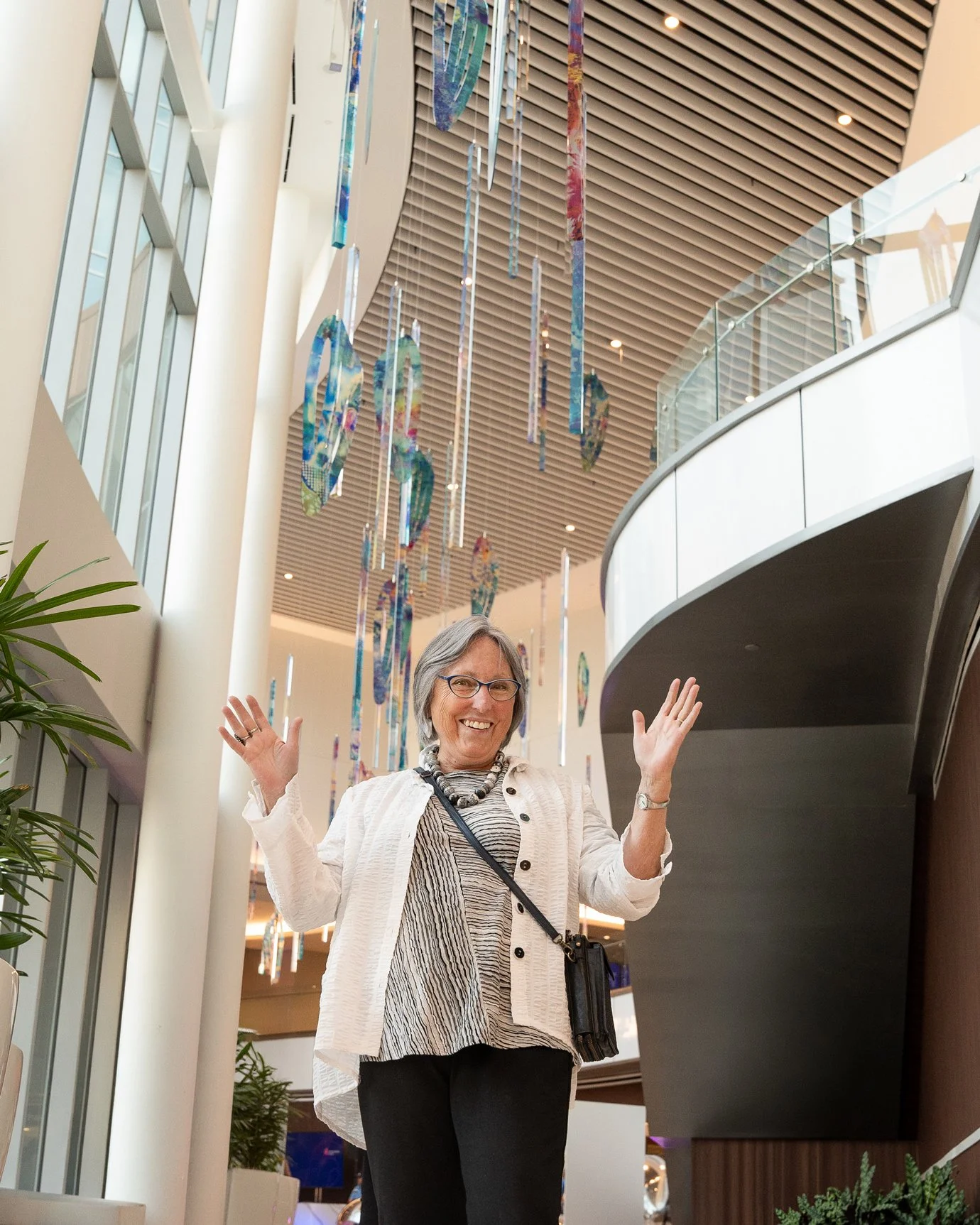 A smiling woman with gray hair, glasses, and a white jacket standing inside a modern, well-lit building with colorful hanging art sculptures overhead.