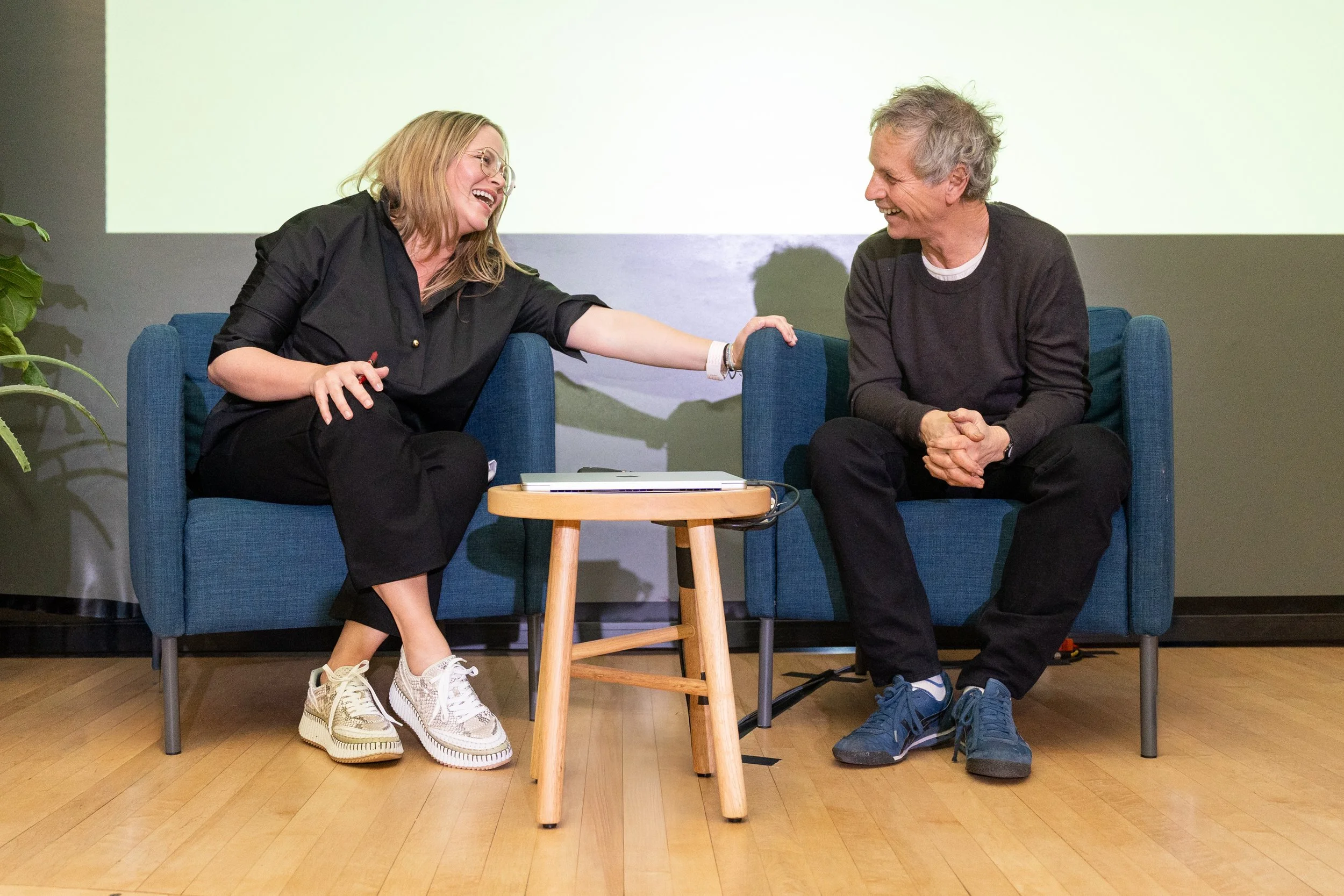 A woman and a man sitting on blue chairs, laughing and sharing a moment of joy, with a small round wooden table between them.