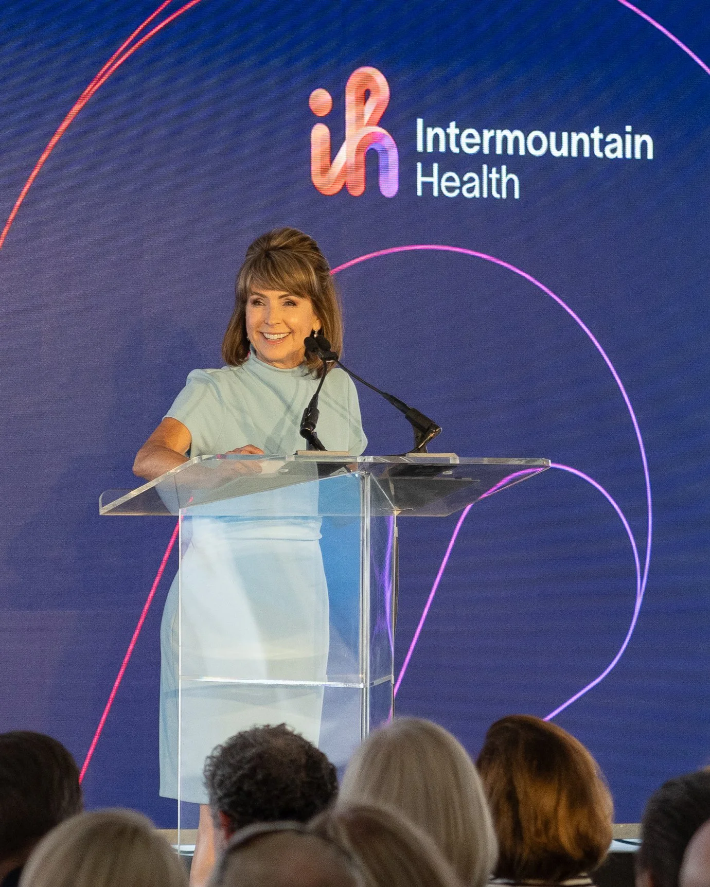 A woman standing at a clear podium with a microphone, smiling, during a presentation or speech at an event hosted by Intermountain Health. A large screen behind her displays the Intermountain Health logo and branding.