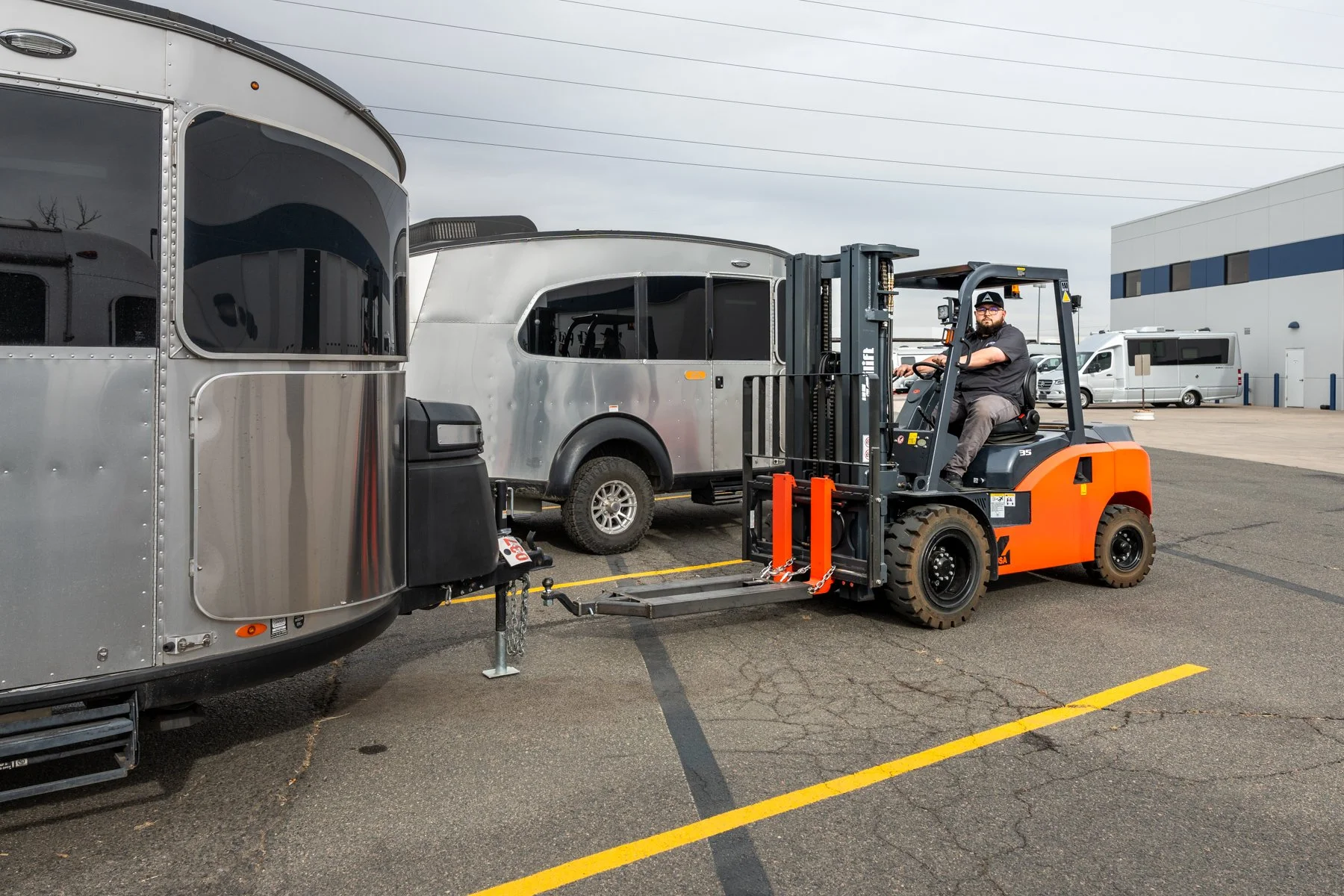 A man operating an orange forklift is loading a silver Airstream trailer in a parking lot, with another silver trailer and a white building in the background.
