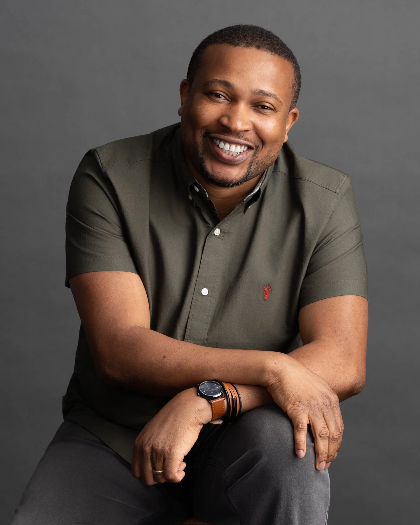 Smiling man with short hair and a beard, wearing a dark green short-sleeve button-up shirt, sitting against a gray background.
