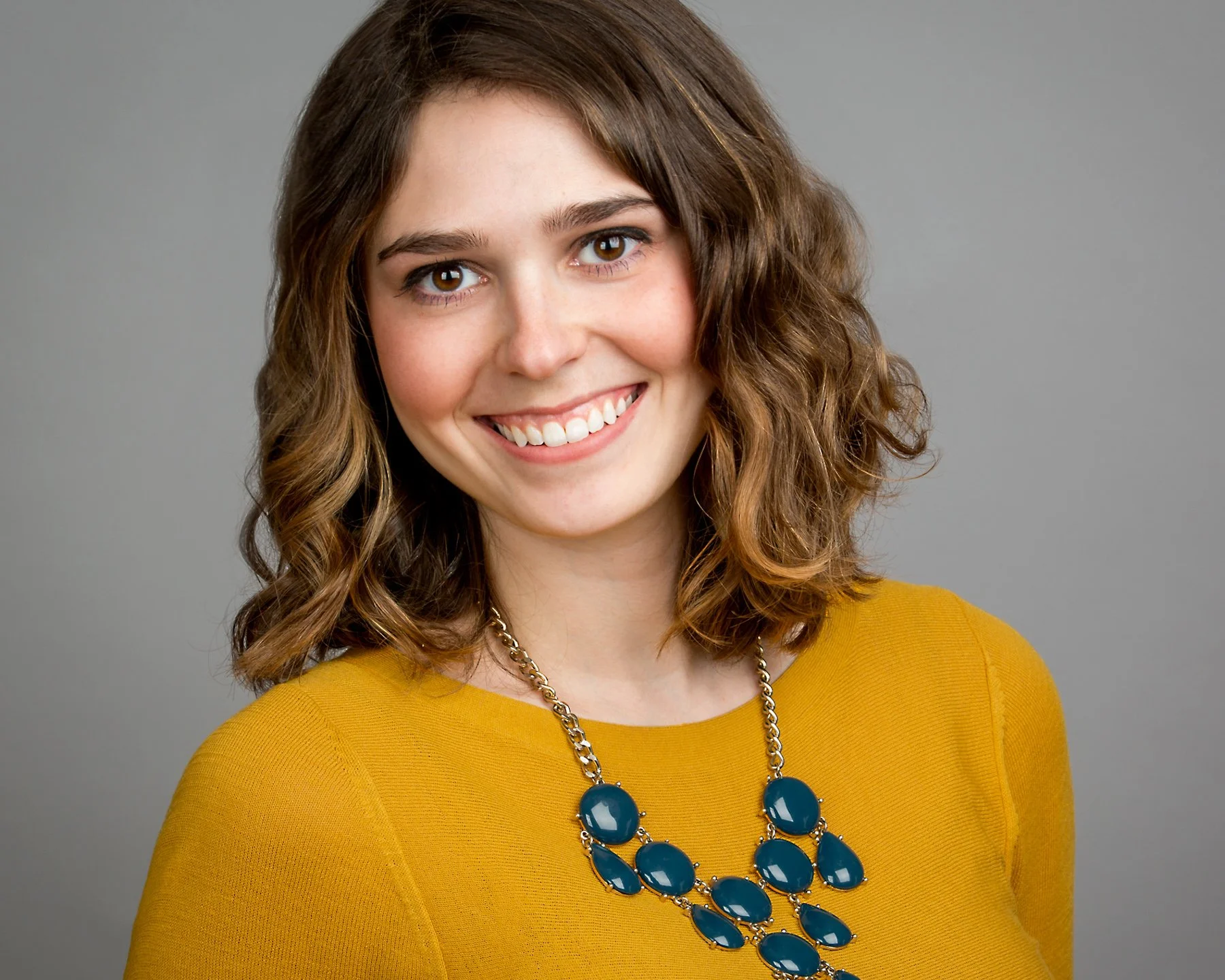 A woman with shoulder-length curly brown hair, wearing a mustard yellow top and a large blue statement necklace, smiling at the camera against a plain grey background.