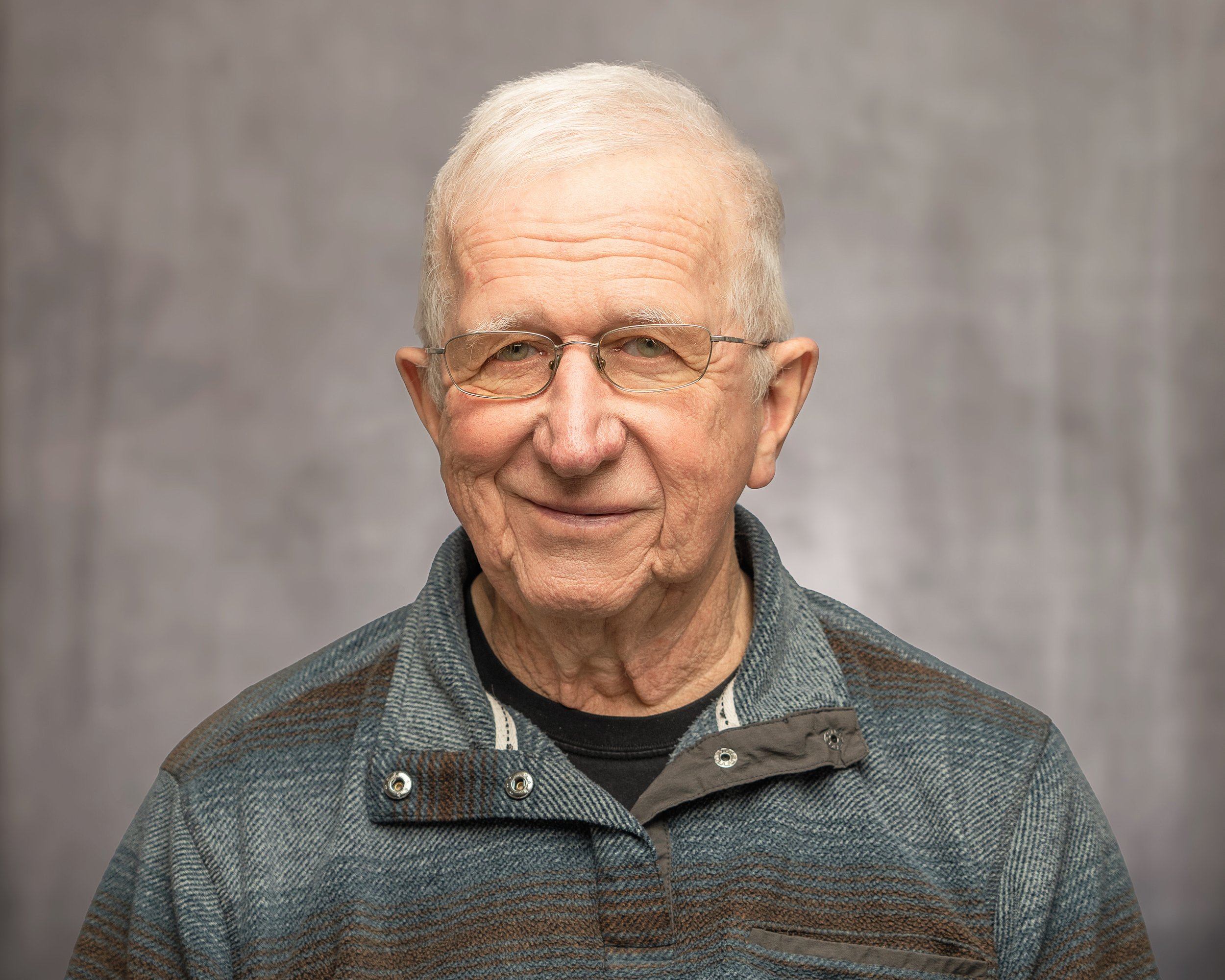 An elderly man with glasses smiling at the camera, wearing a plaid sweater with a collar, standing against a plain gray background.
