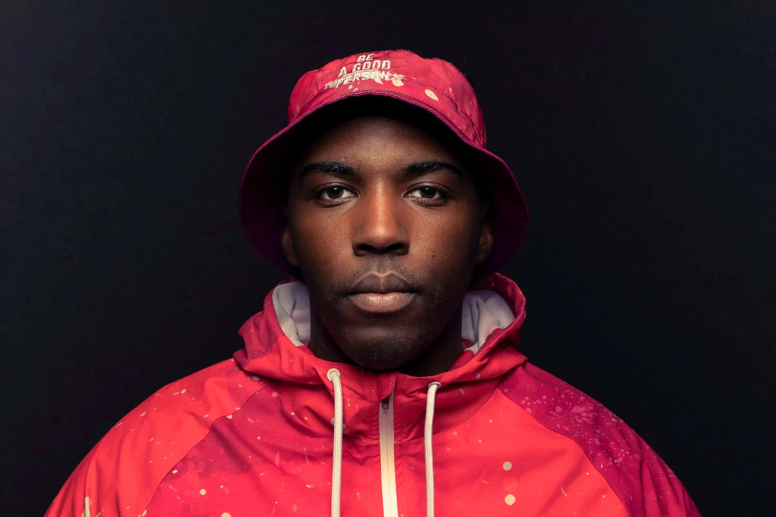 A young man wearing a red jacket and a red bucket hat against a dark background.