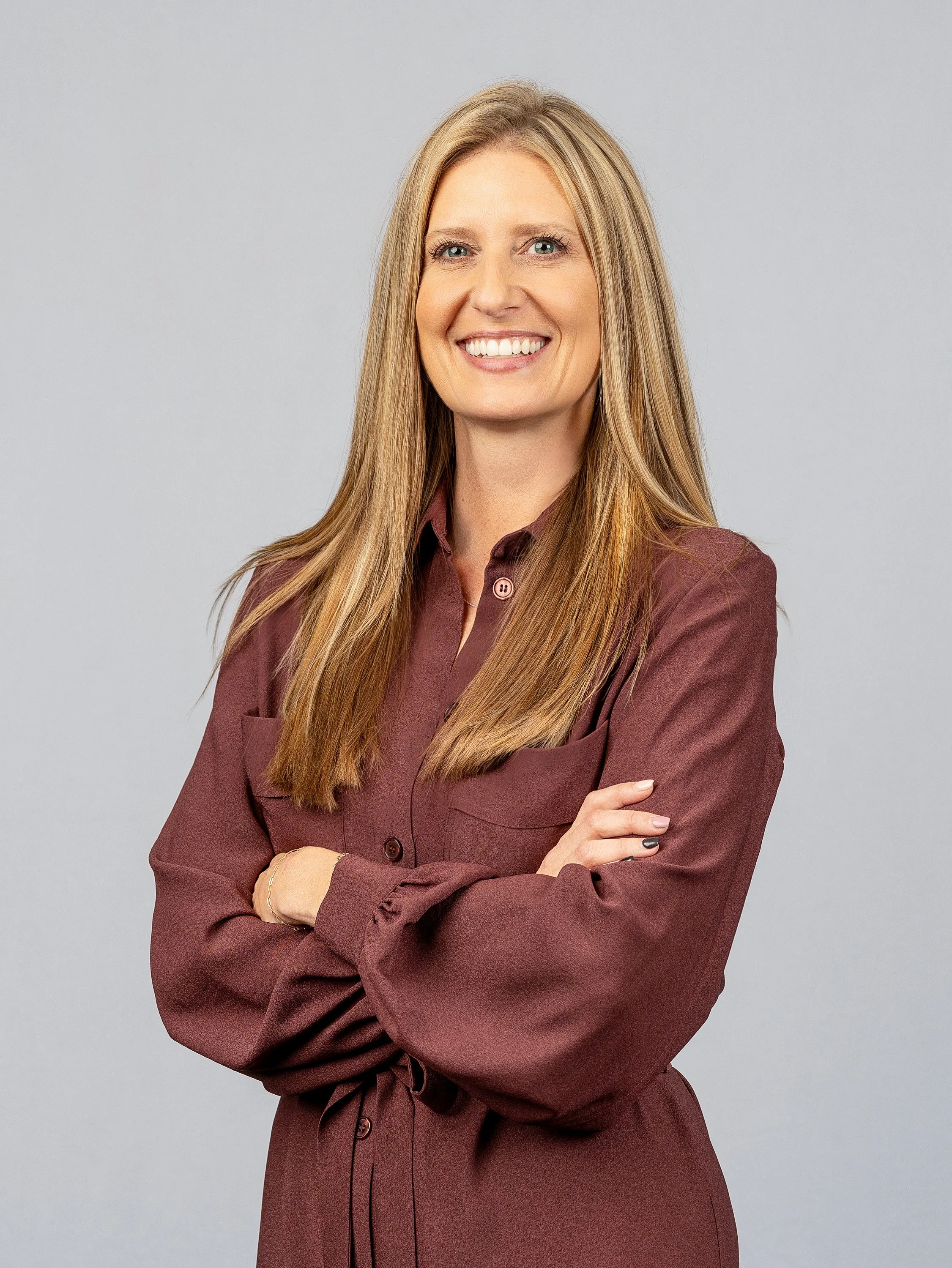 A woman with long, straight blonde hair smiling with arms crossed, wearing a maroon button-up shirt against a light gray background.