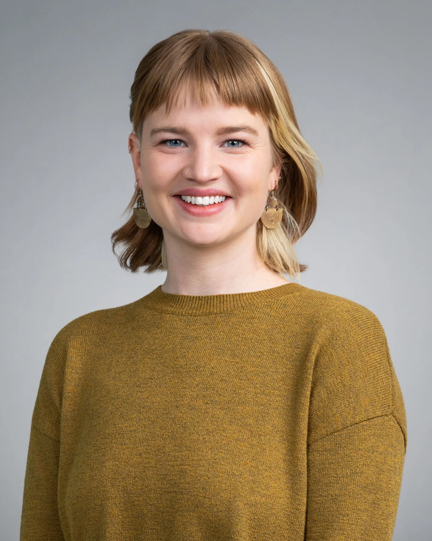 A smiling woman with short red hair wearing a mustard-colored sweater and heart-shaped earrings, standing against a gray background.