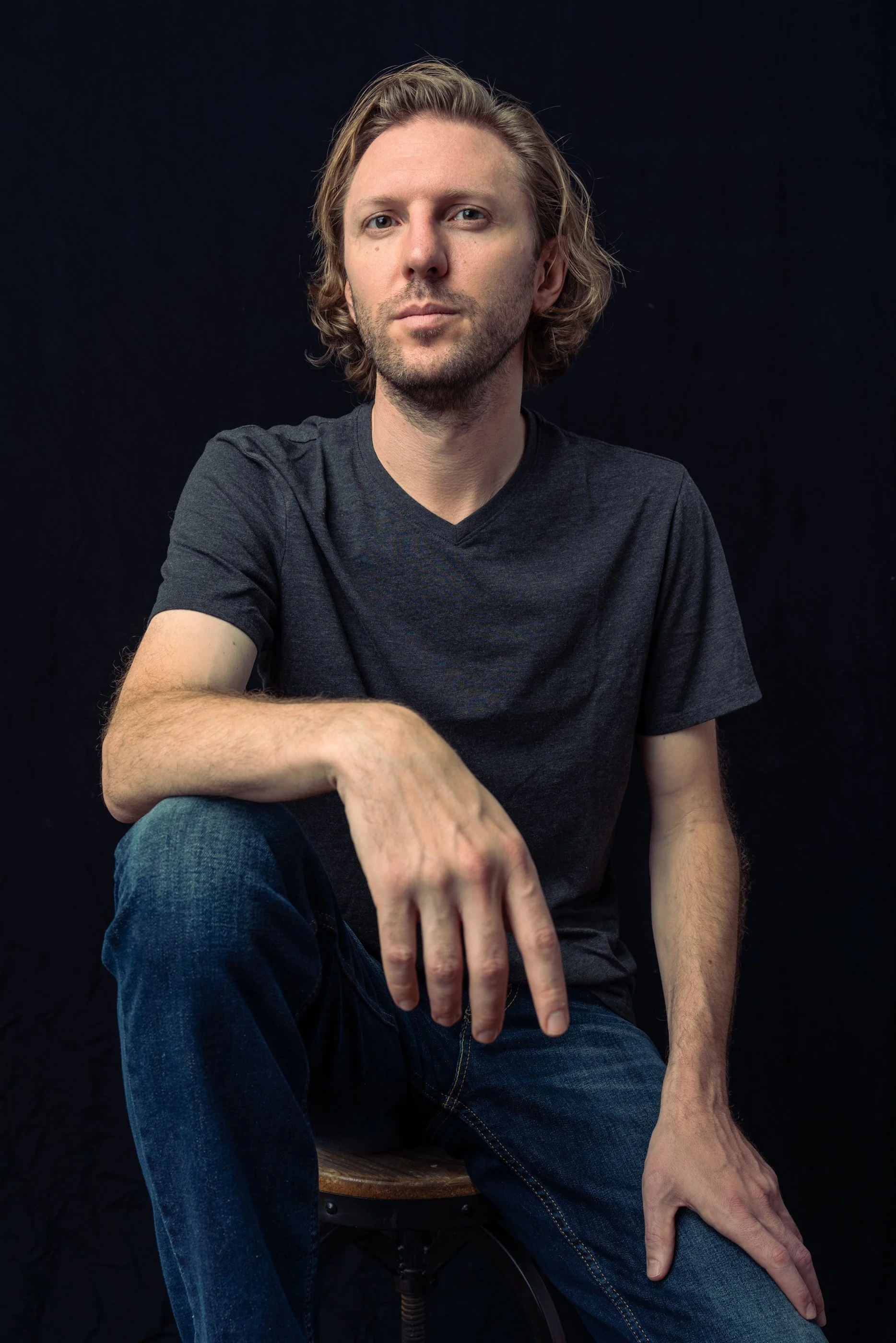 A man with shoulder-length blonde hair, wearing a black t-shirt, seated on a wooden stool against a black background.
