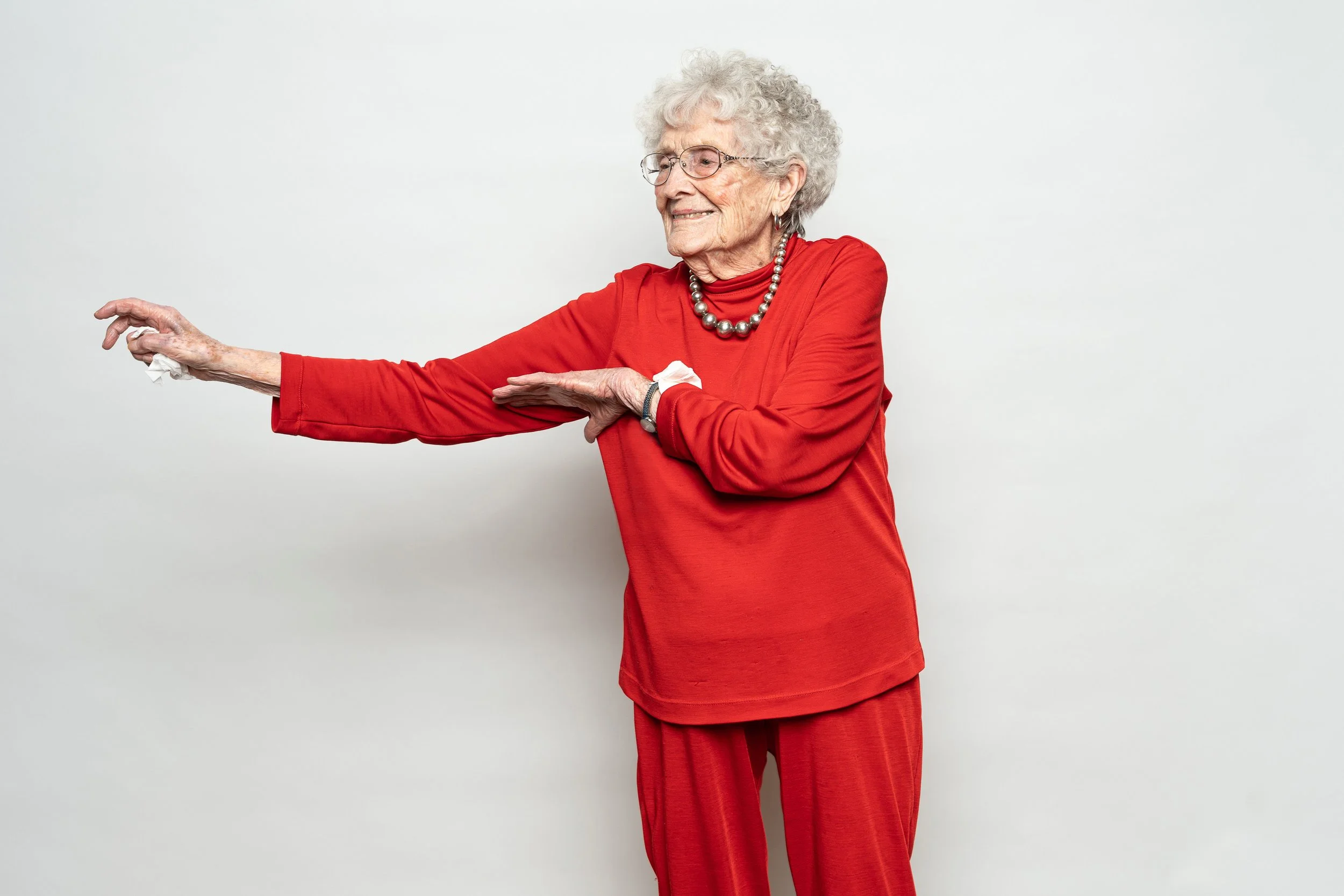 An elderly woman with curly gray hair, glasses, and a pearl necklace is wearing a red outfit and stretching her arms against a plain white background.