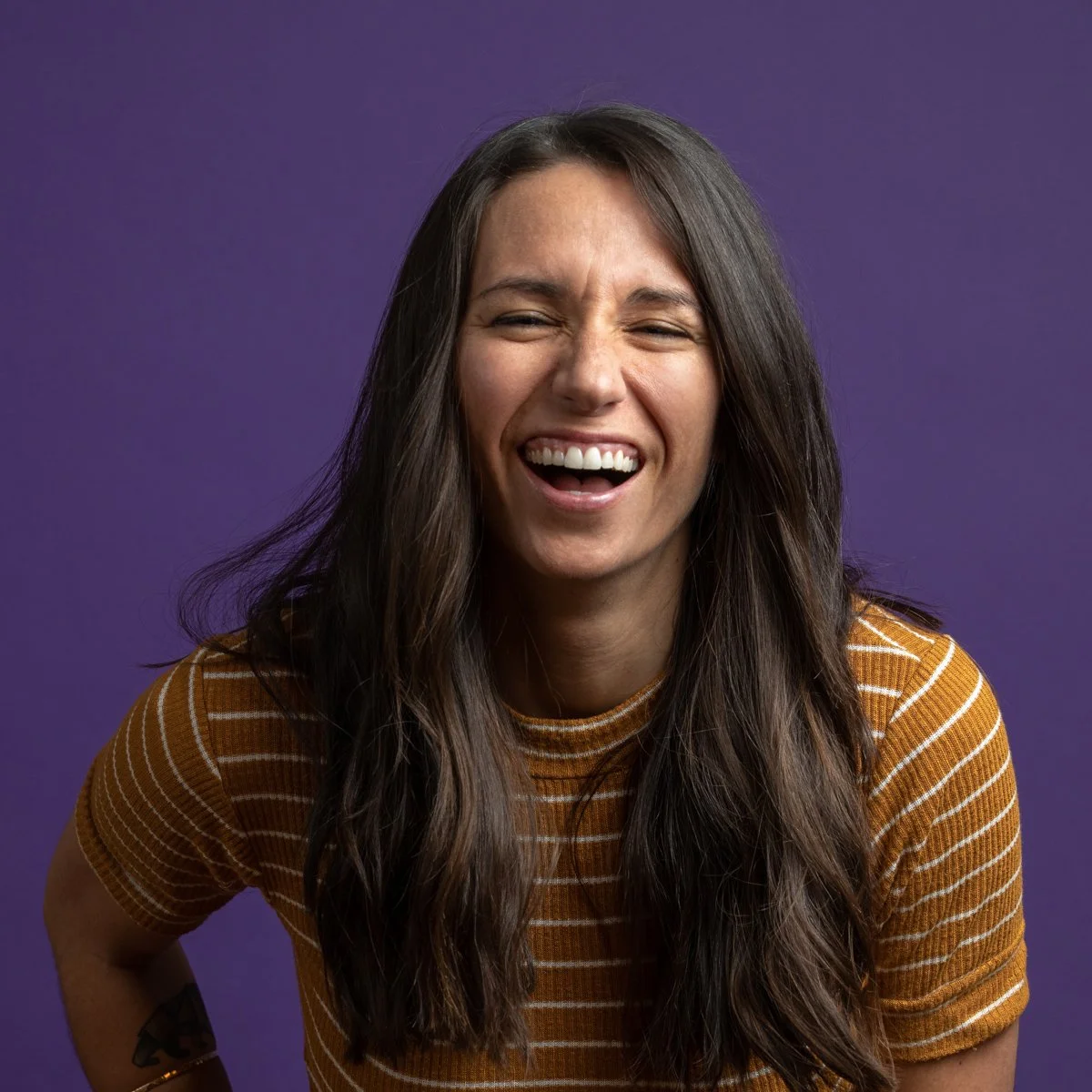 A woman with long dark hair is smiling and squinting in front of a purple background, wearing a brown striped shirt.