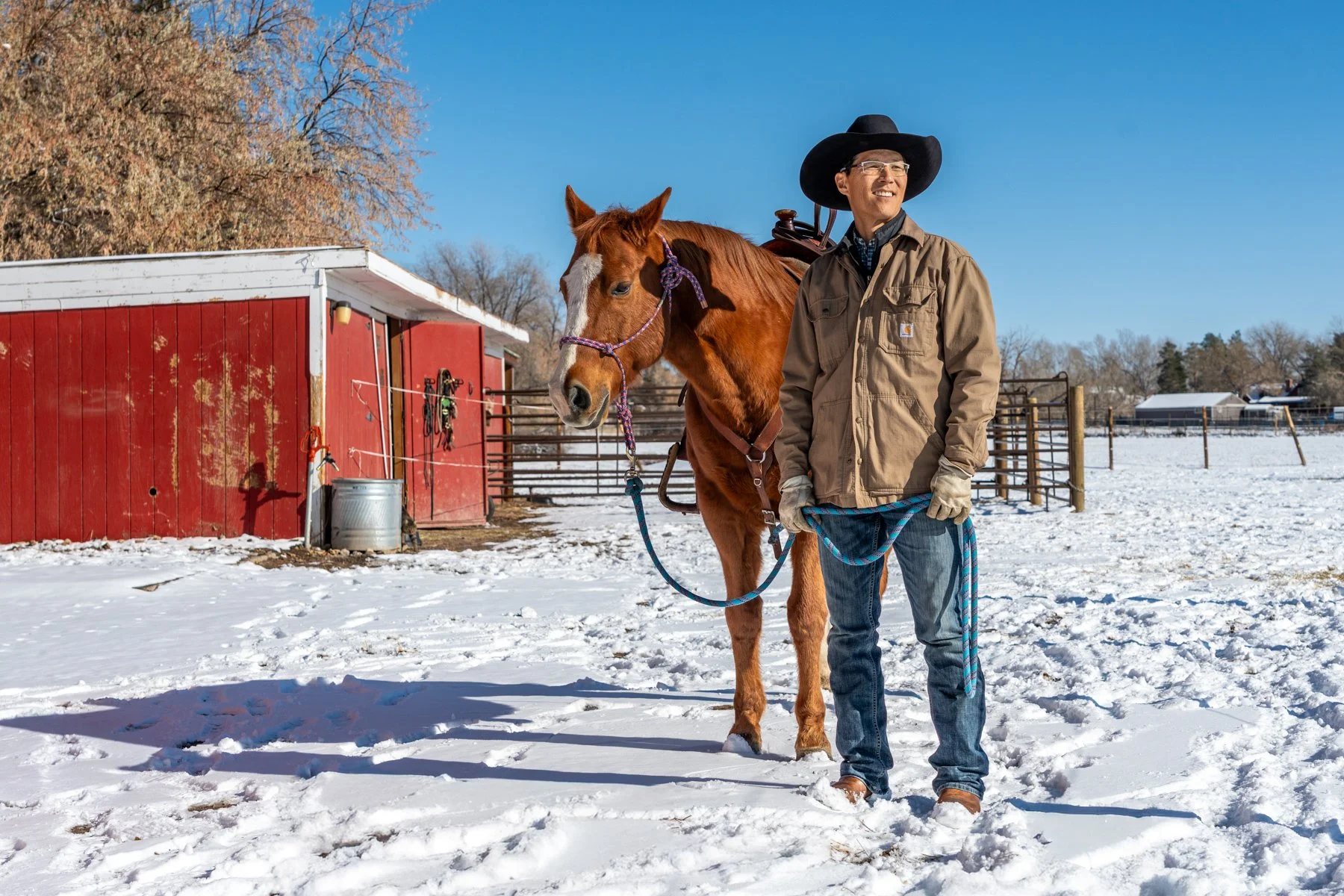 A man in winter clothing and a black cowboy hat stands next to a brown horse with a purple halter, in a snowy farmyard with a red barn in the background.