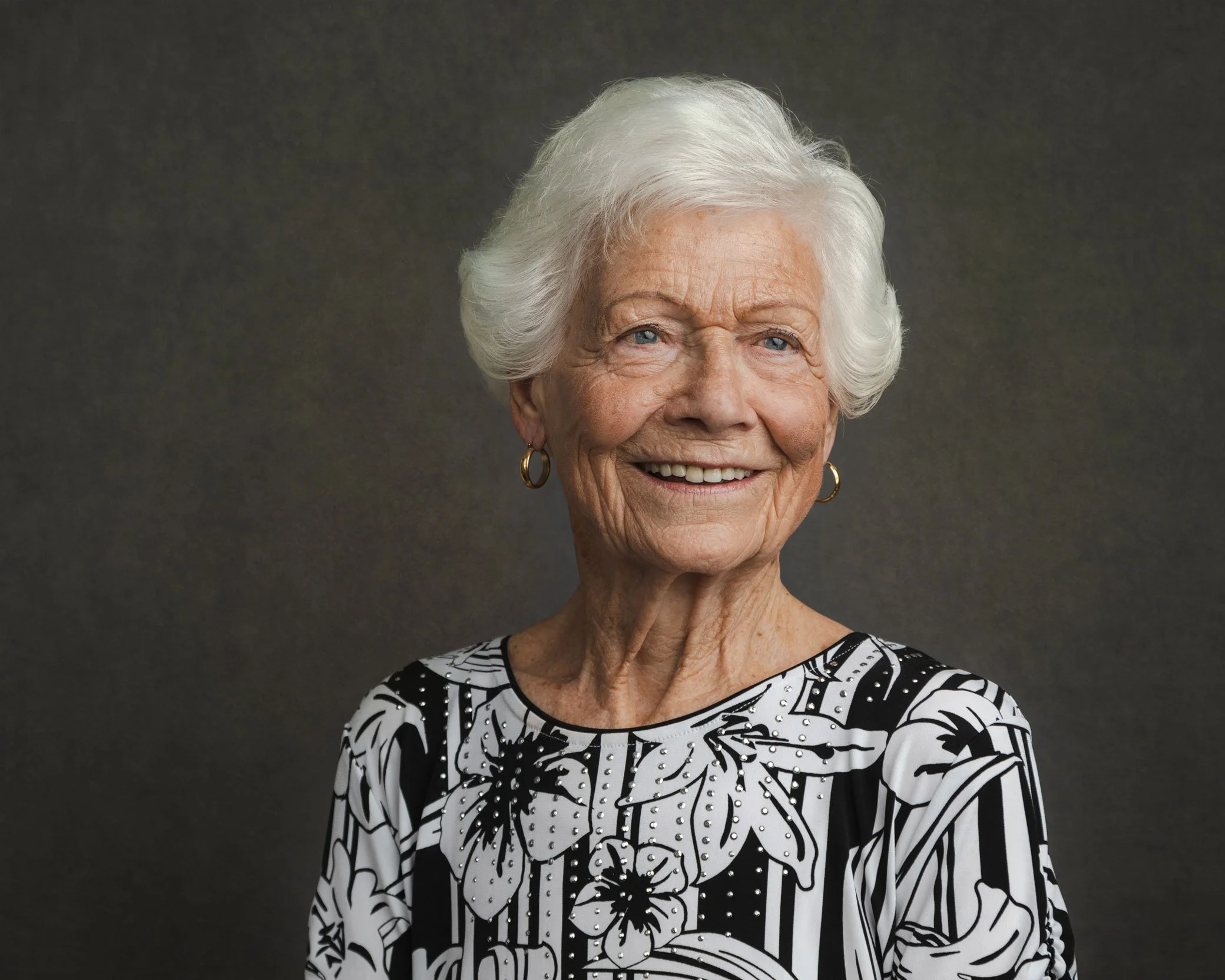 An elderly woman with white hair, smiling, wearing a black and white floral patterned blouse with small pearl or rhinestone accents, standing against a dark gray background.
