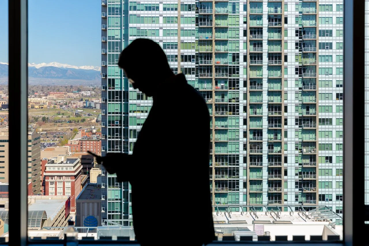 Silhouette of a man looking down at his phone, standing by a large window in a high-rise building, with cityscape and mountains in the background.