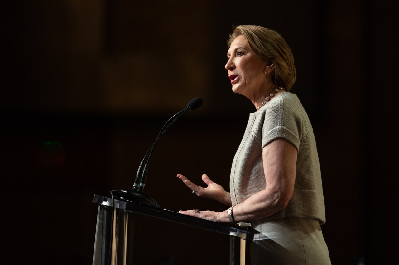 Carly Fiorina giving a speech at a podium with a microphone, wearing a beige outfit and a pearl necklace, against a dark background in Denver, CO