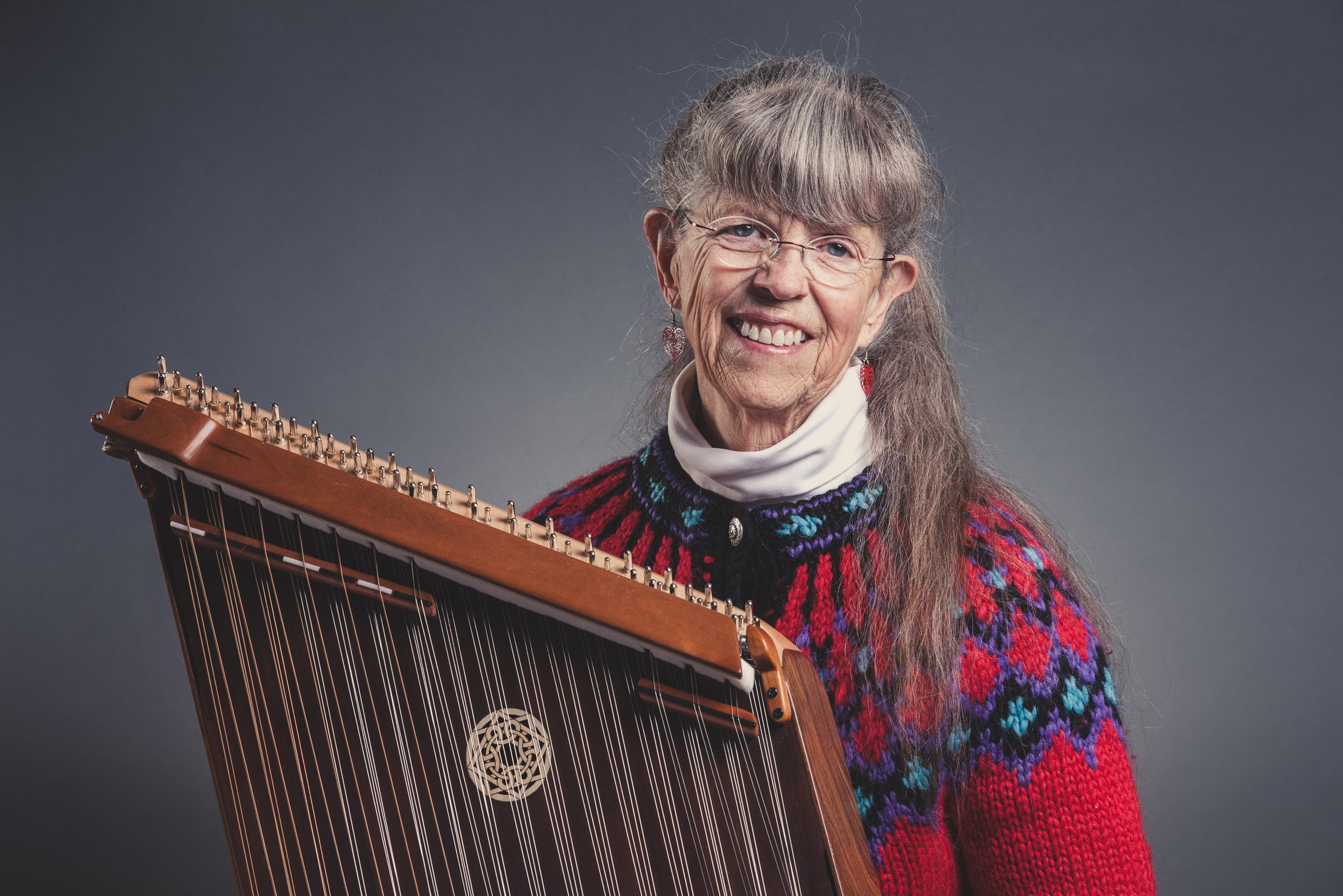 An elderly woman with glasses and long gray hair, wearing a colorful knit sweater and earrings, smiling while holding a hammered dulcimer against a gray background.