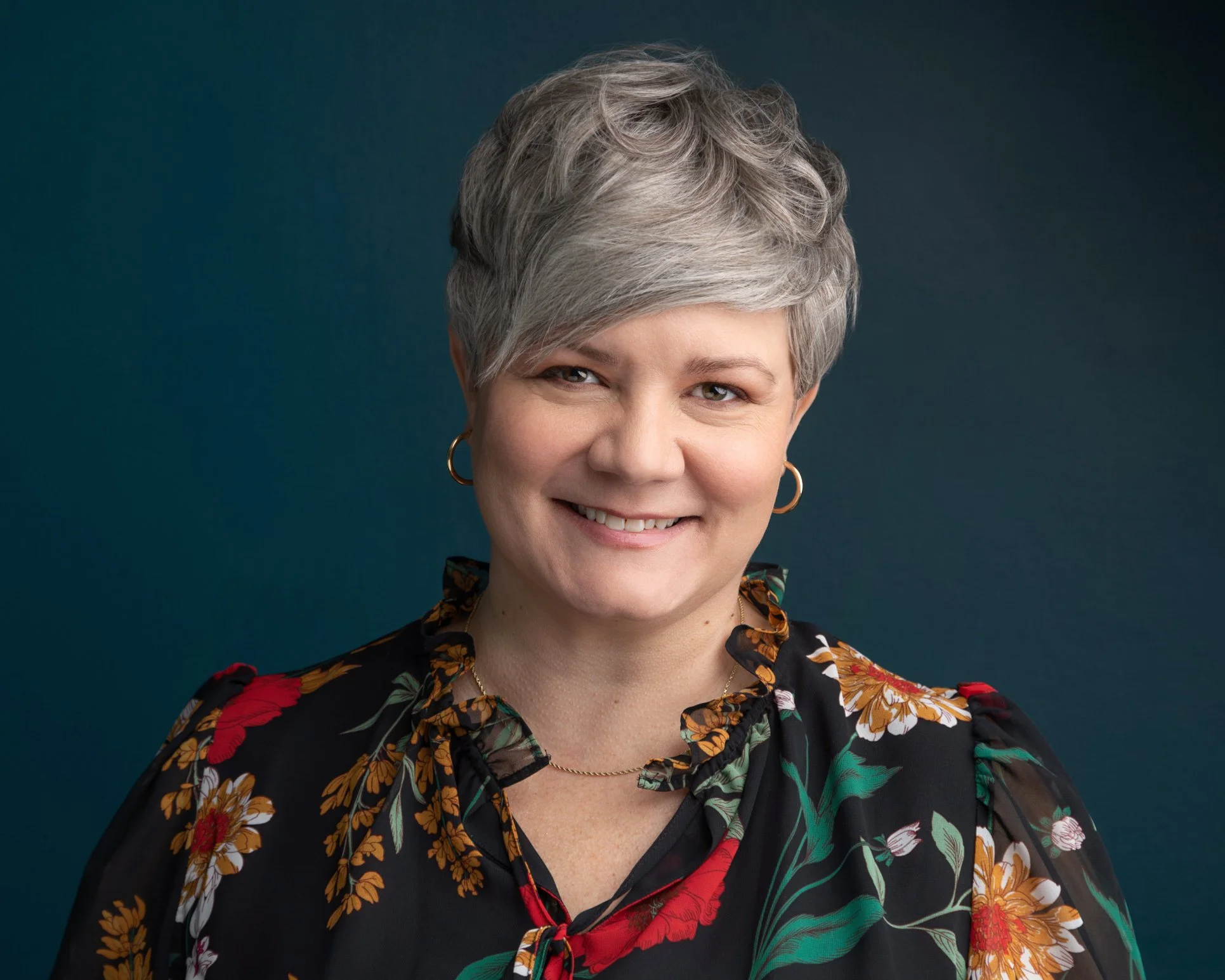 A mature woman with short gray hair, wearing a black floral blouse with red, yellow, and green flowers, gold hoop earrings, and a subtle smile, standing against a dark teal background.