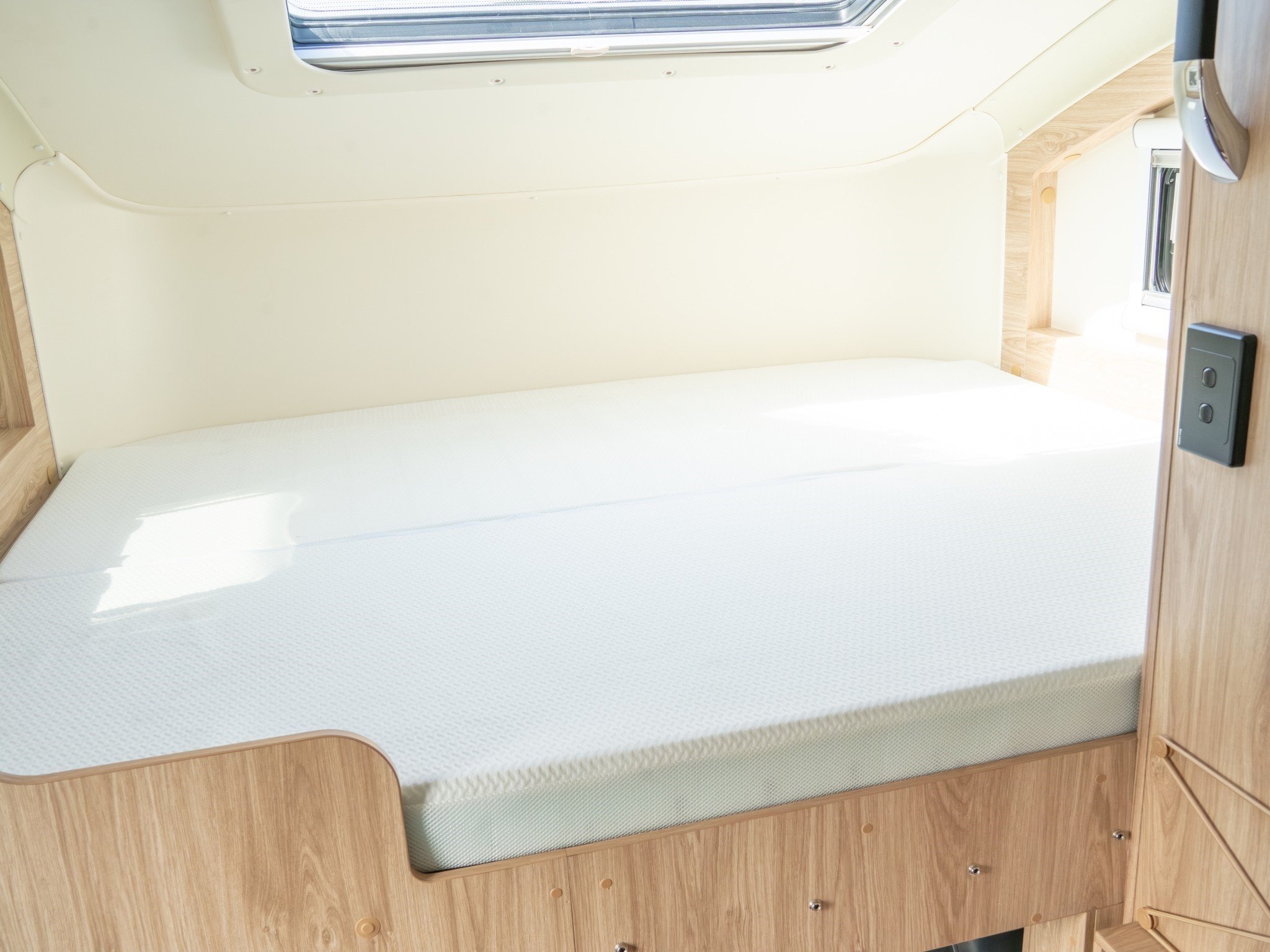 A bedsit with a neatly made mattress on a wooden frame, with a slide-out compartment underneath. There is a window at the head of the bed, with a ceiling hatch above, and a wooden side table to the right.