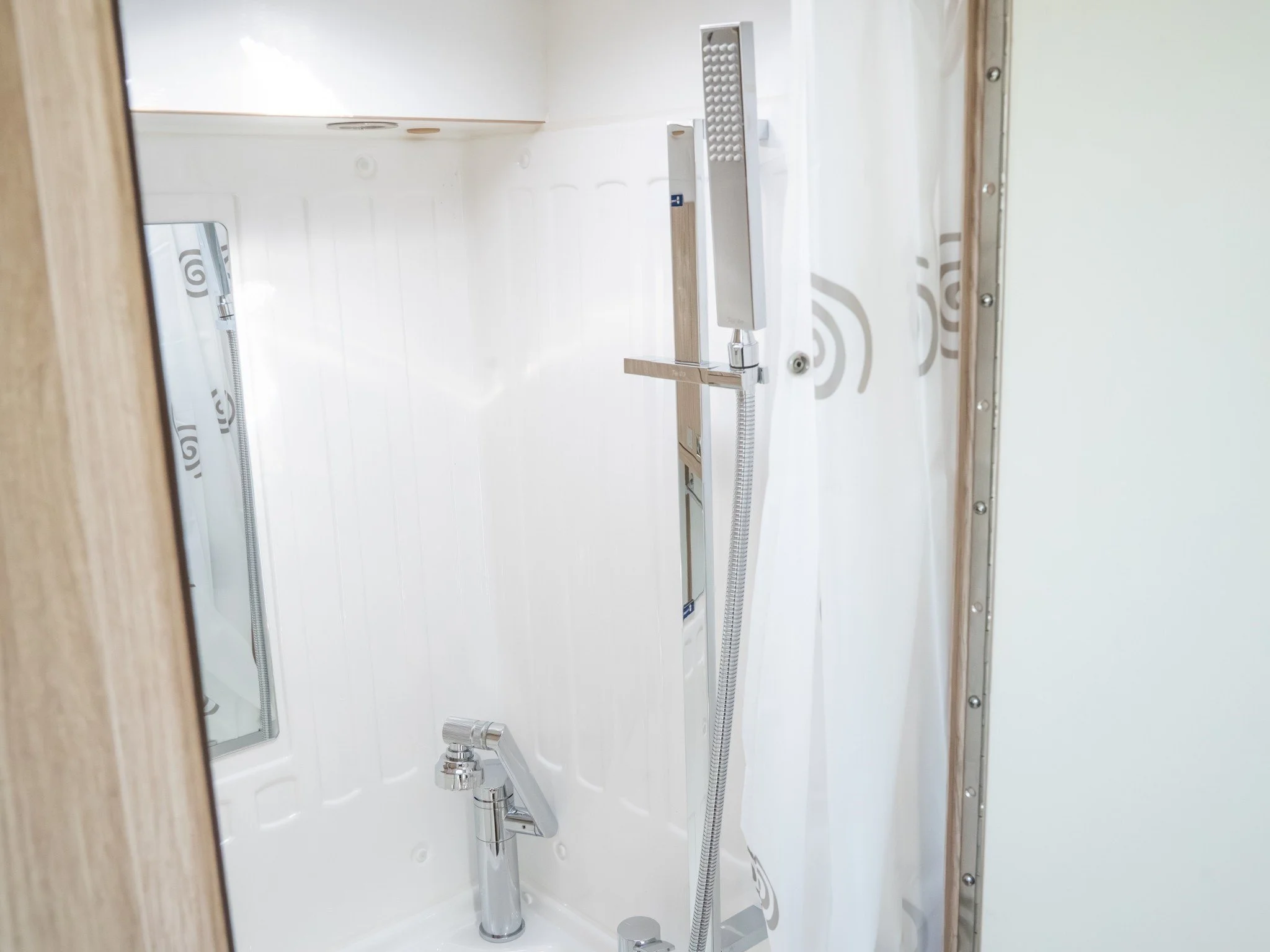 Interior of a shower with white walls, a handheld showerhead attached to a vertical bar, and a mirror on the left side.