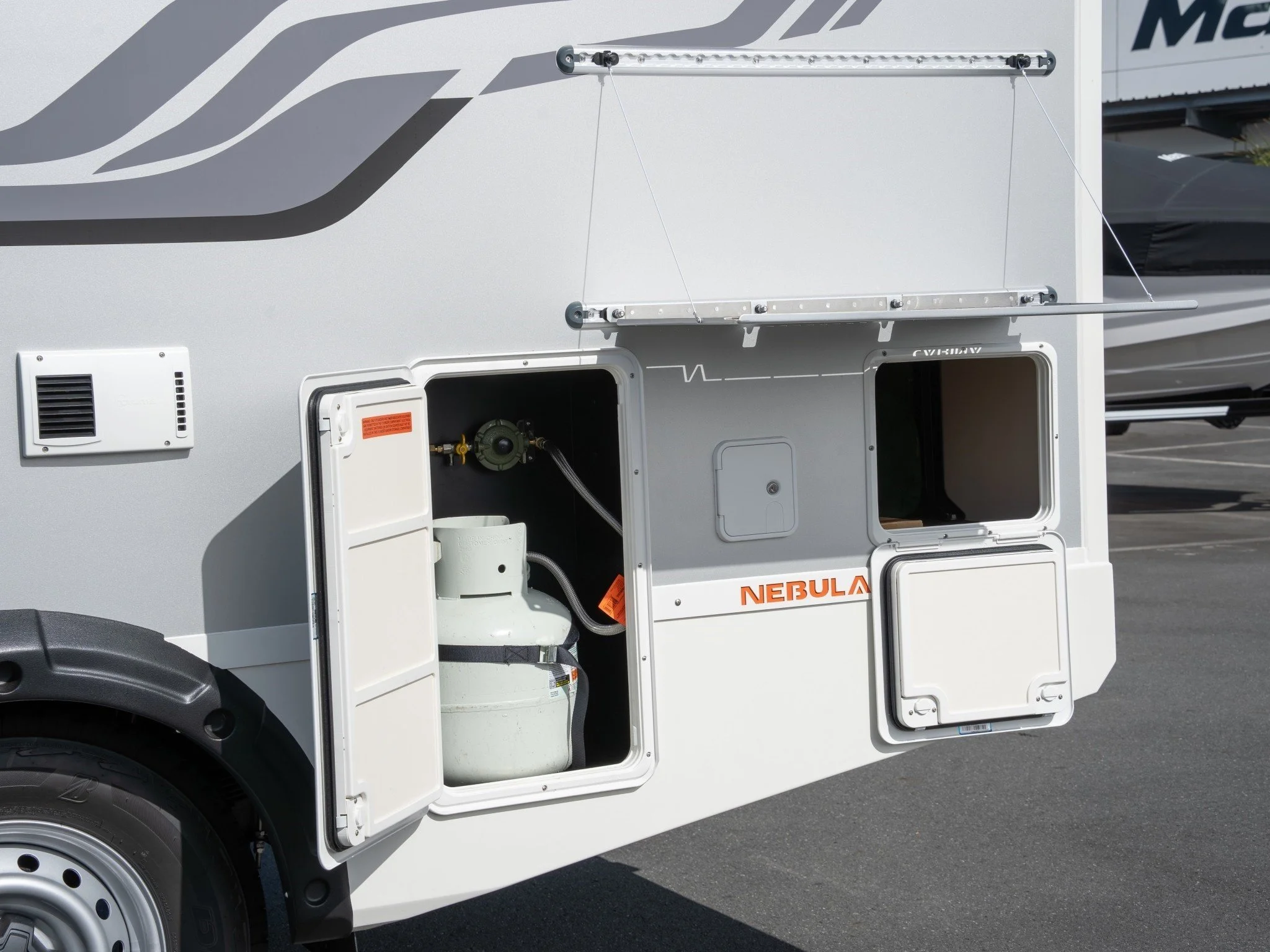 A section of a white RV with open service doors, revealing storage compartments and utility connections. The RV has a graphic design and the word Nebula written on it.