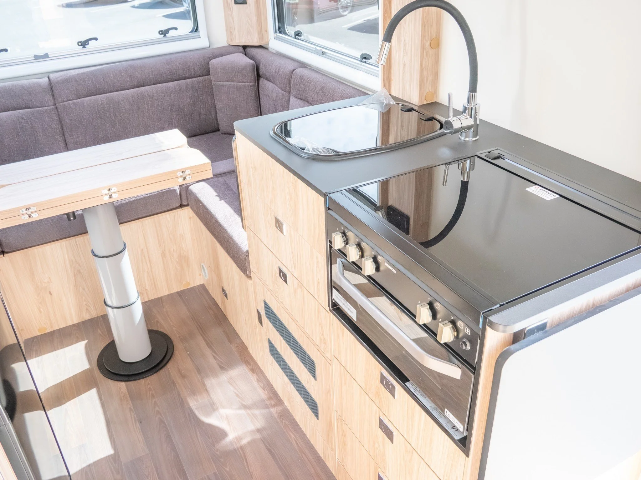 Interior of a camper van kitchen with a small stove, sink, wood cabinetry, a gray sofa, and windows showing outside parking.